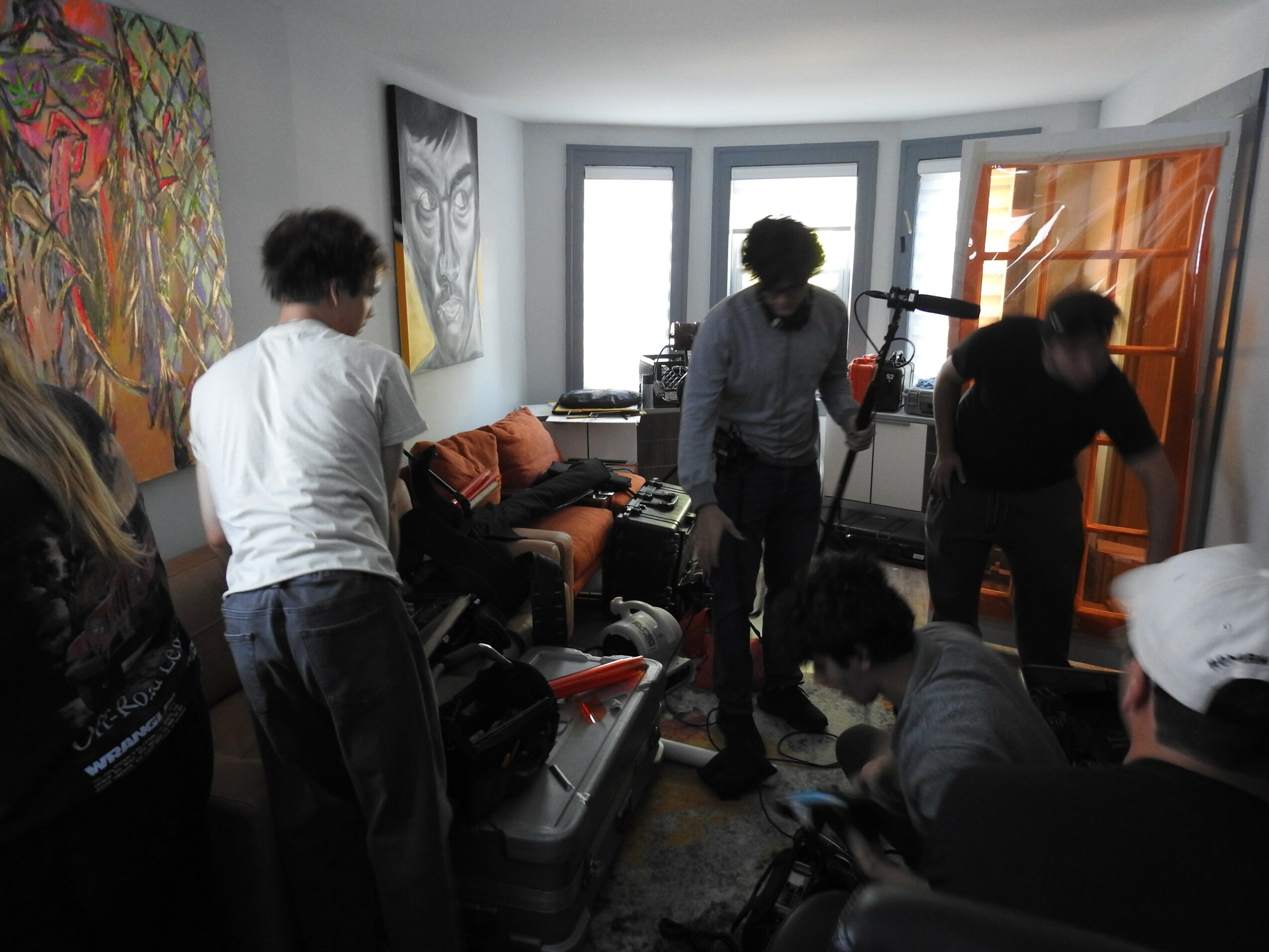 Group of people in a living room, some standing and some sitting, with a window and artwork on the wall.