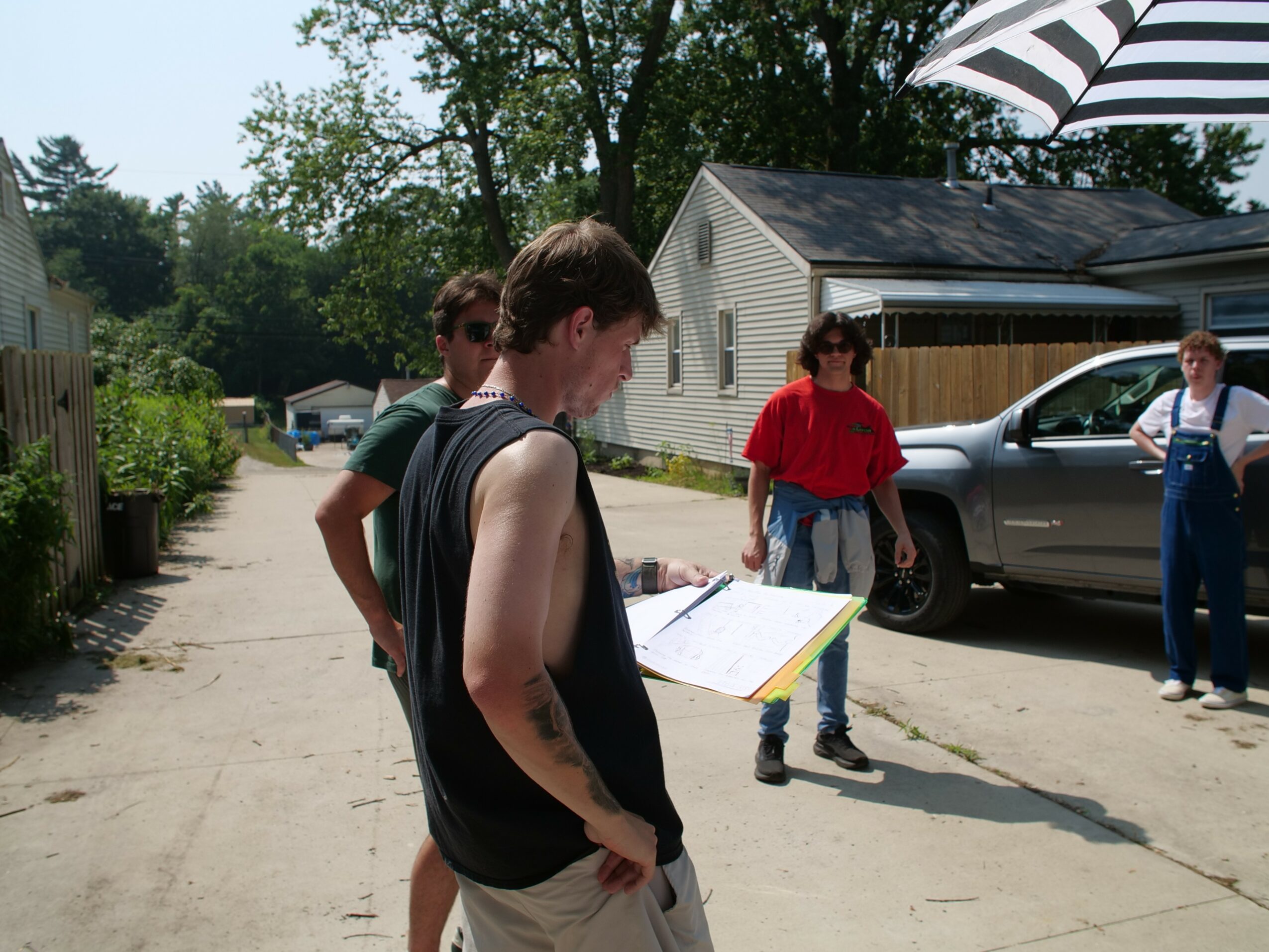 Group of five people standing outdoors on a sunny day, near a house and parked car, engaged in conversation.