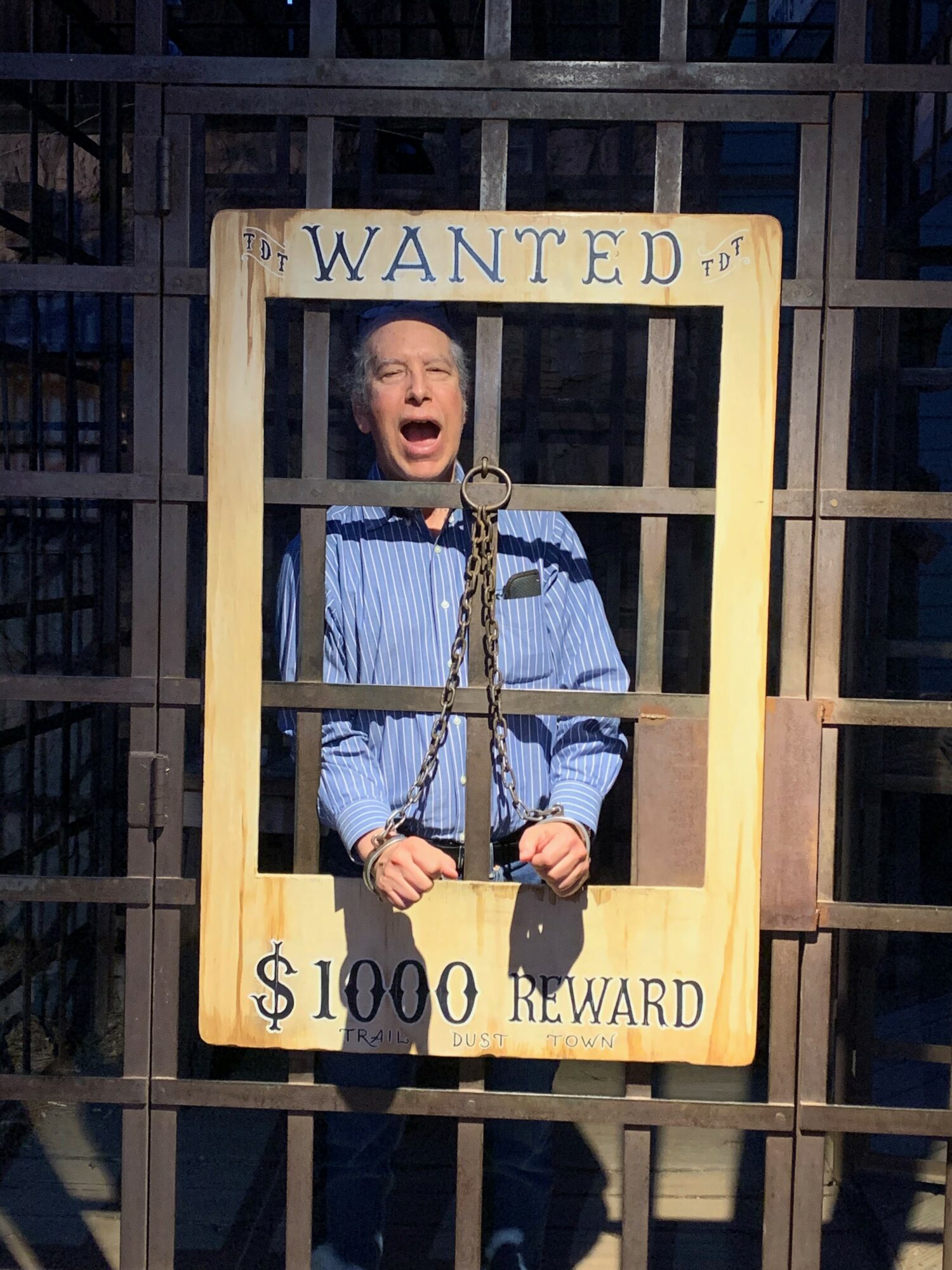Person with mouth open behind a wanted poster frame, inside a cage or bars, with a sign reading 'WANTED' and reward amount.