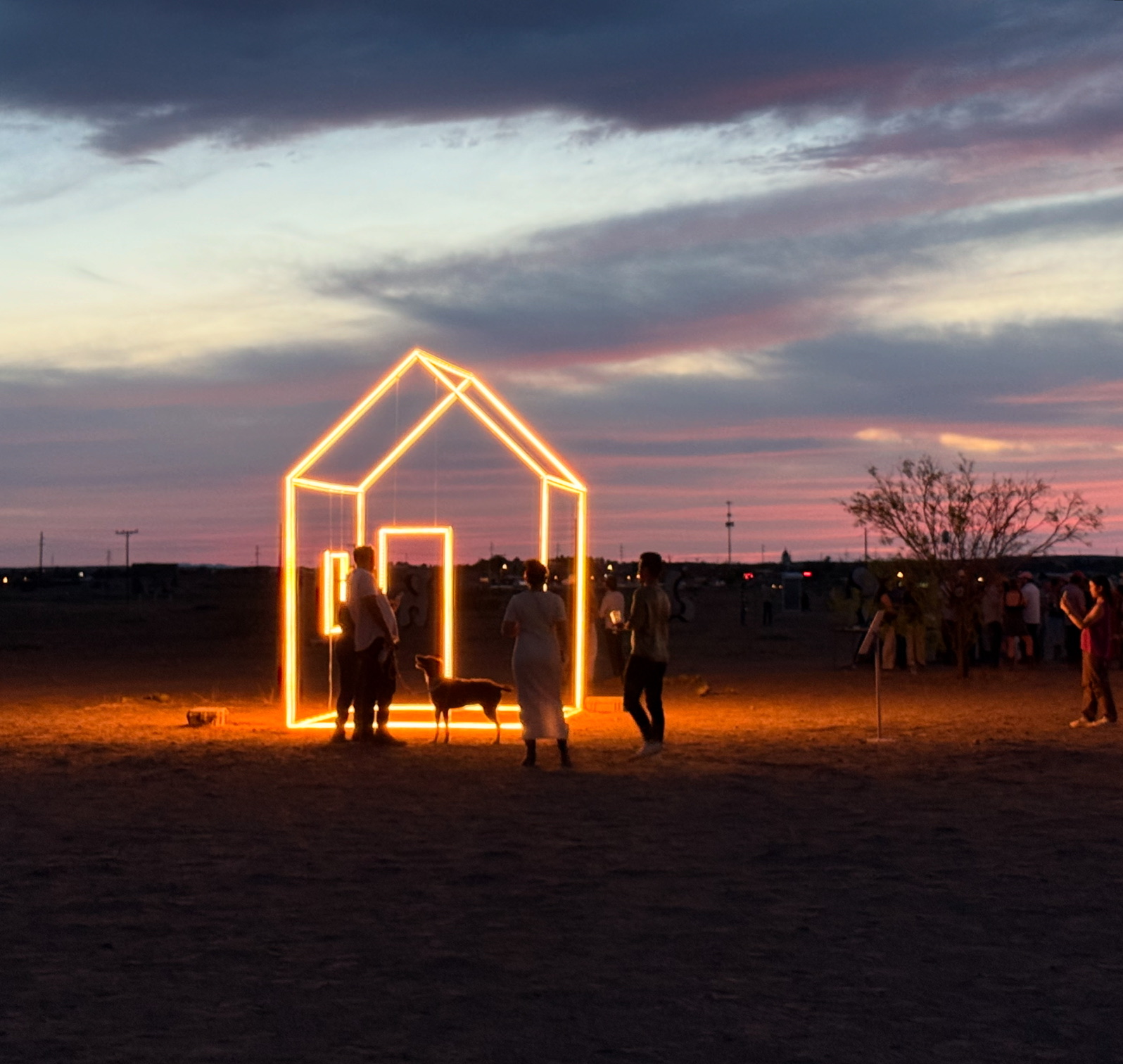 People gather around illuminated house-shaped structure at sunset in open field.