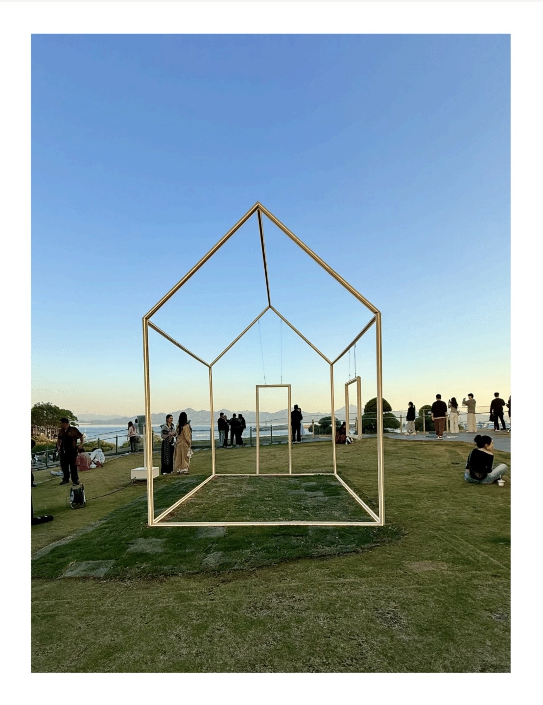 Open metal frame structure resembling a house on a grassy area with people and a body of water in the background.