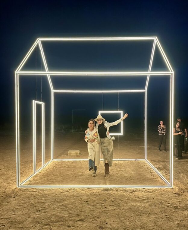 People walking through a glowing geometric light installation on a dirt surface at night.