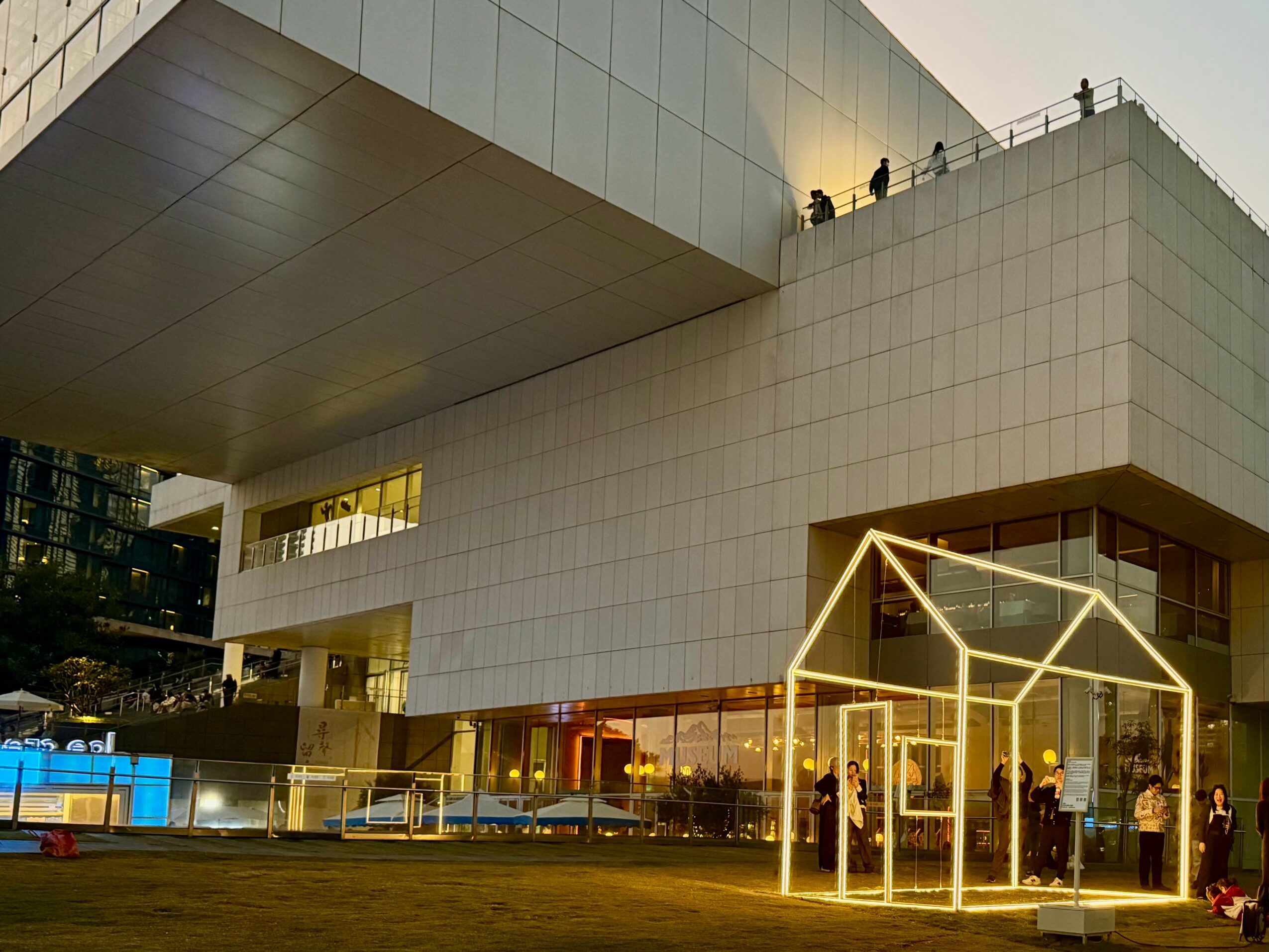 Modern building with large overhang, glass entrance, and people on rooftop and ground level.