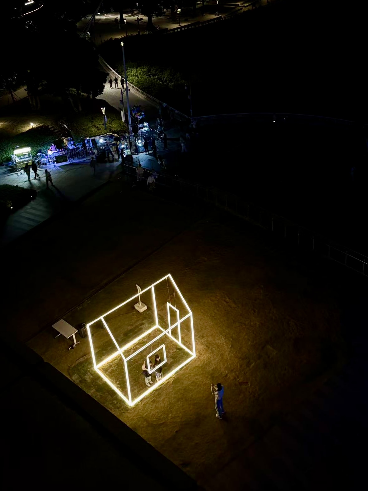 Night scene with illuminated house-shaped structure and a person nearby on a grassy area.