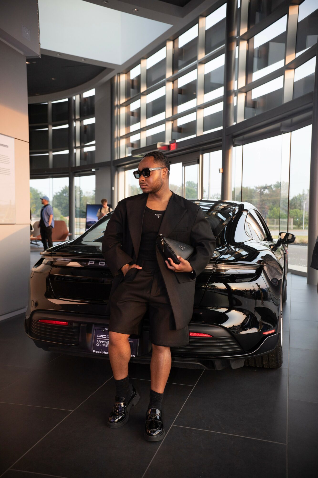 Person in black outfit and sunglasses standing in front of a black car inside a modern building with large windows.