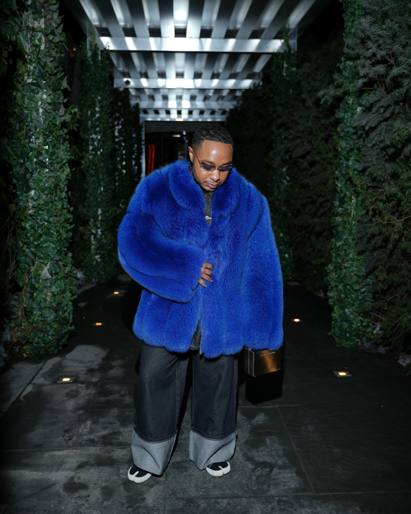 Person wearing a large blue fur coat, glasses, and dark pants walking in a corridor with greenery walls.