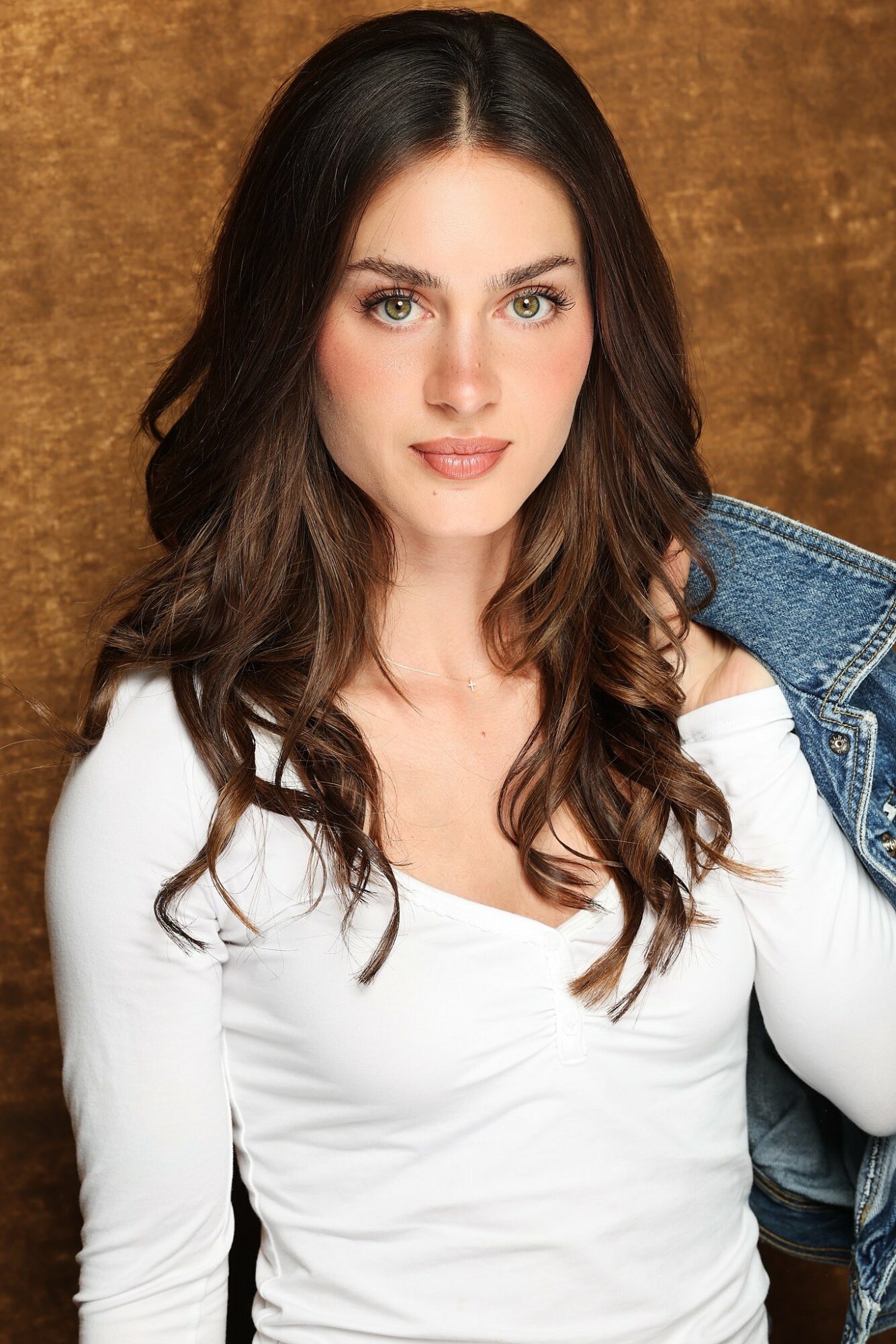 Young woman with long dark hair, wearing a white top and denim jacket, against a brown background.