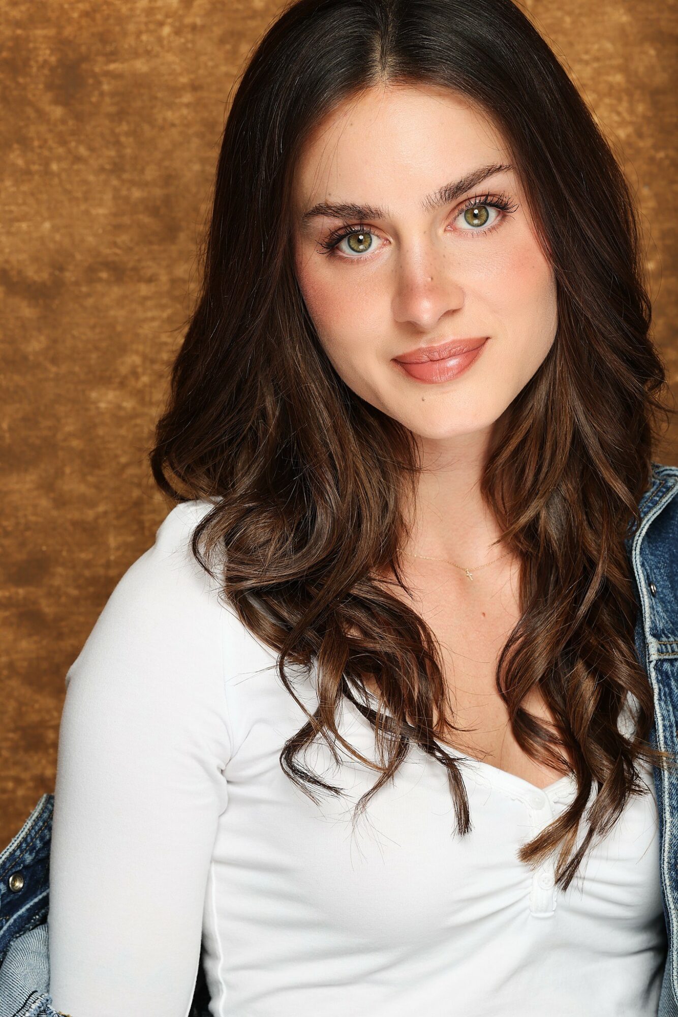 Young woman with long wavy brown hair, green eyes, wearing a white top, smiling, against a brown background.