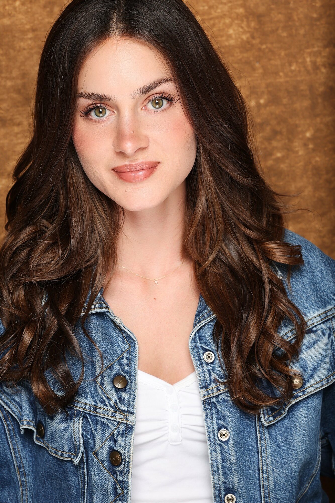 Young woman with long wavy brown hair, wearing a denim jacket and white top, smiling softly.