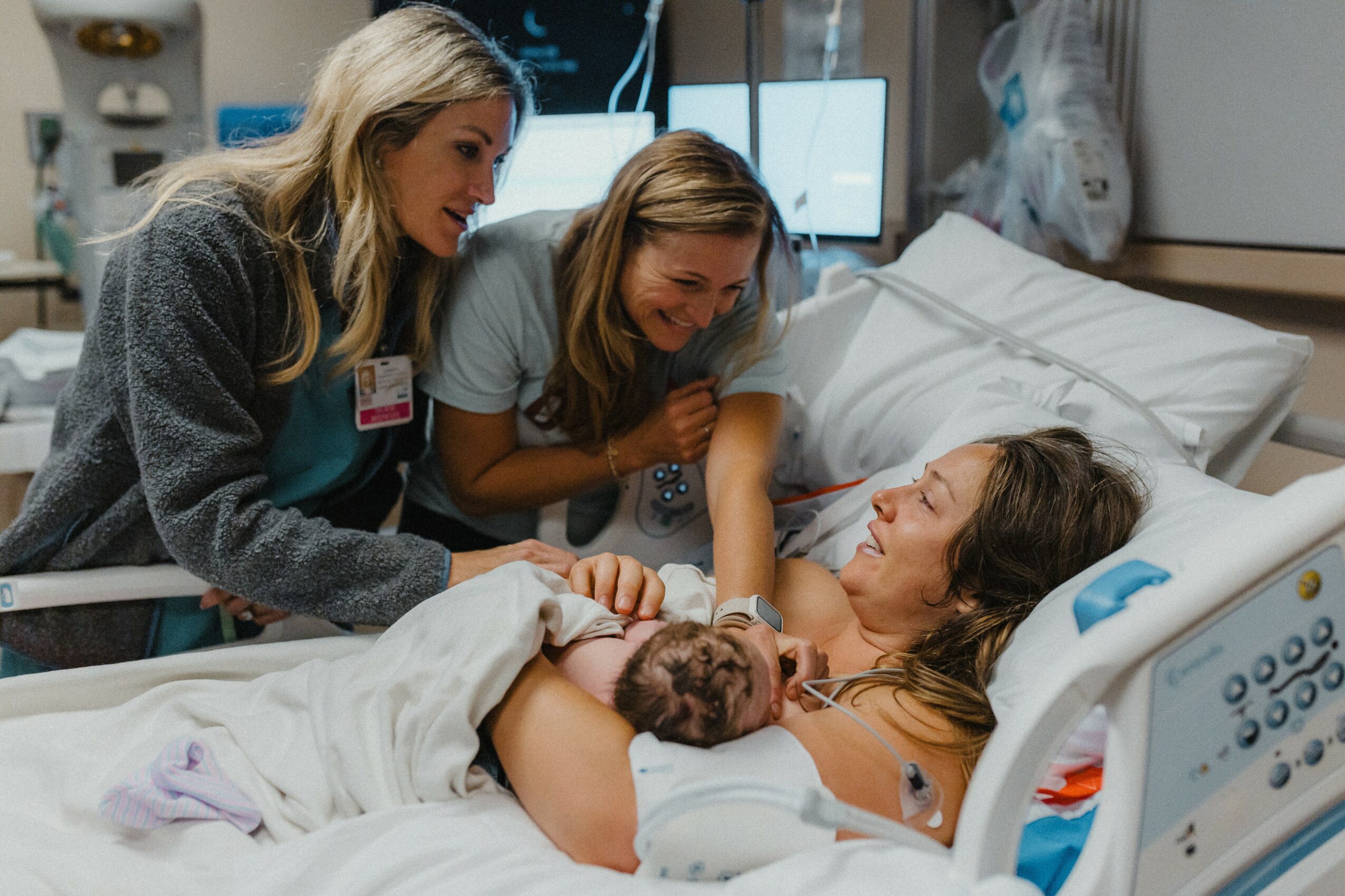 Woman in hospital bed holding newborn with two women beside her, in a hospital room.