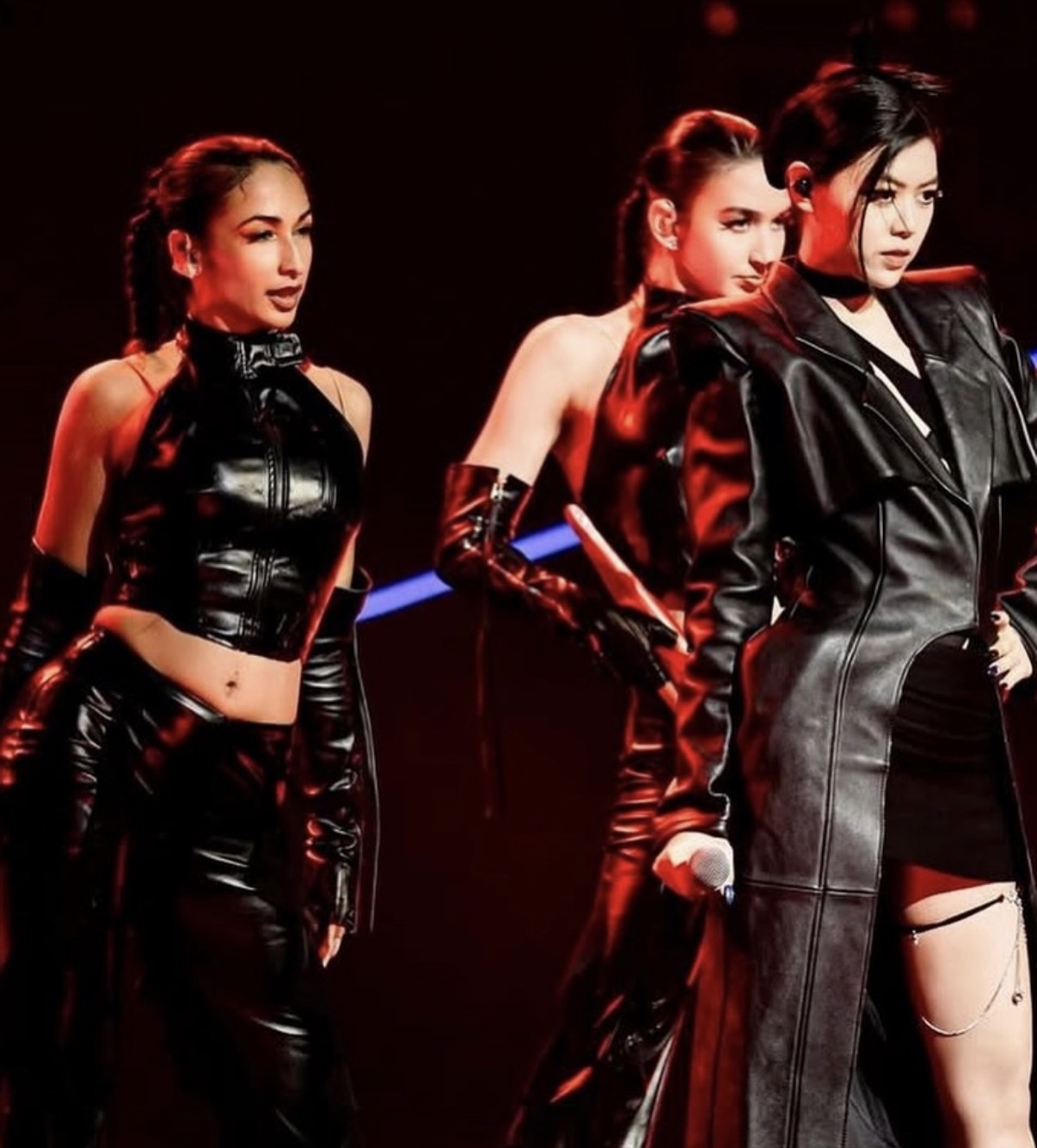Three women in black outfits on stage with dark background, one with a torn dress, all with serious expressions.