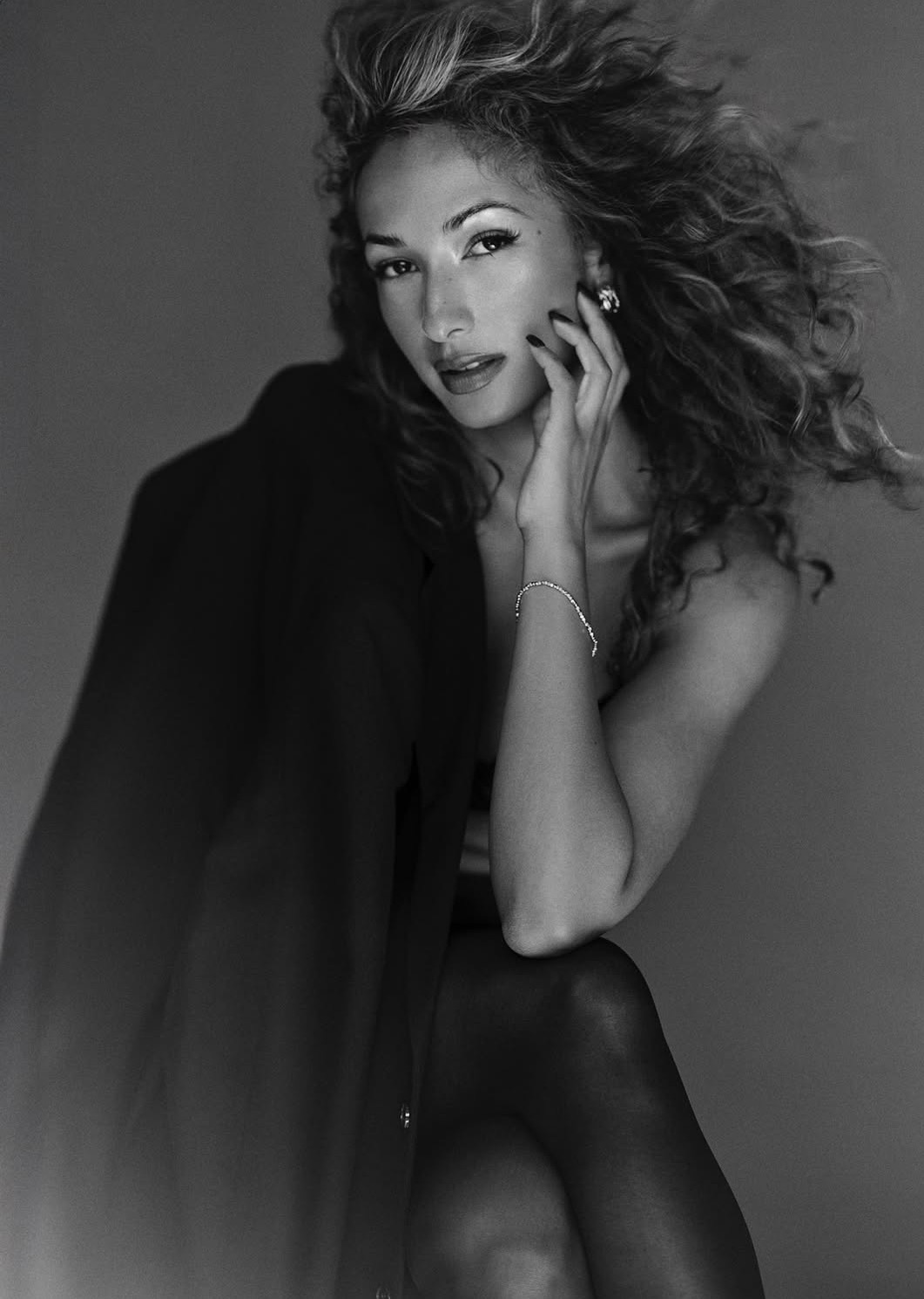Woman with curly hair, sitting and touching her face, wearing dark clothing, in black and white.