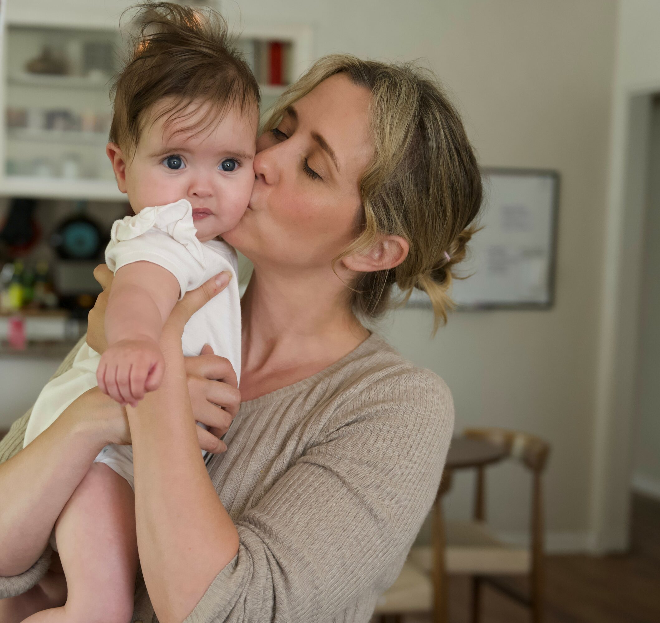 Woman holding a baby close, kissing the baby's cheek in a cozy room.
