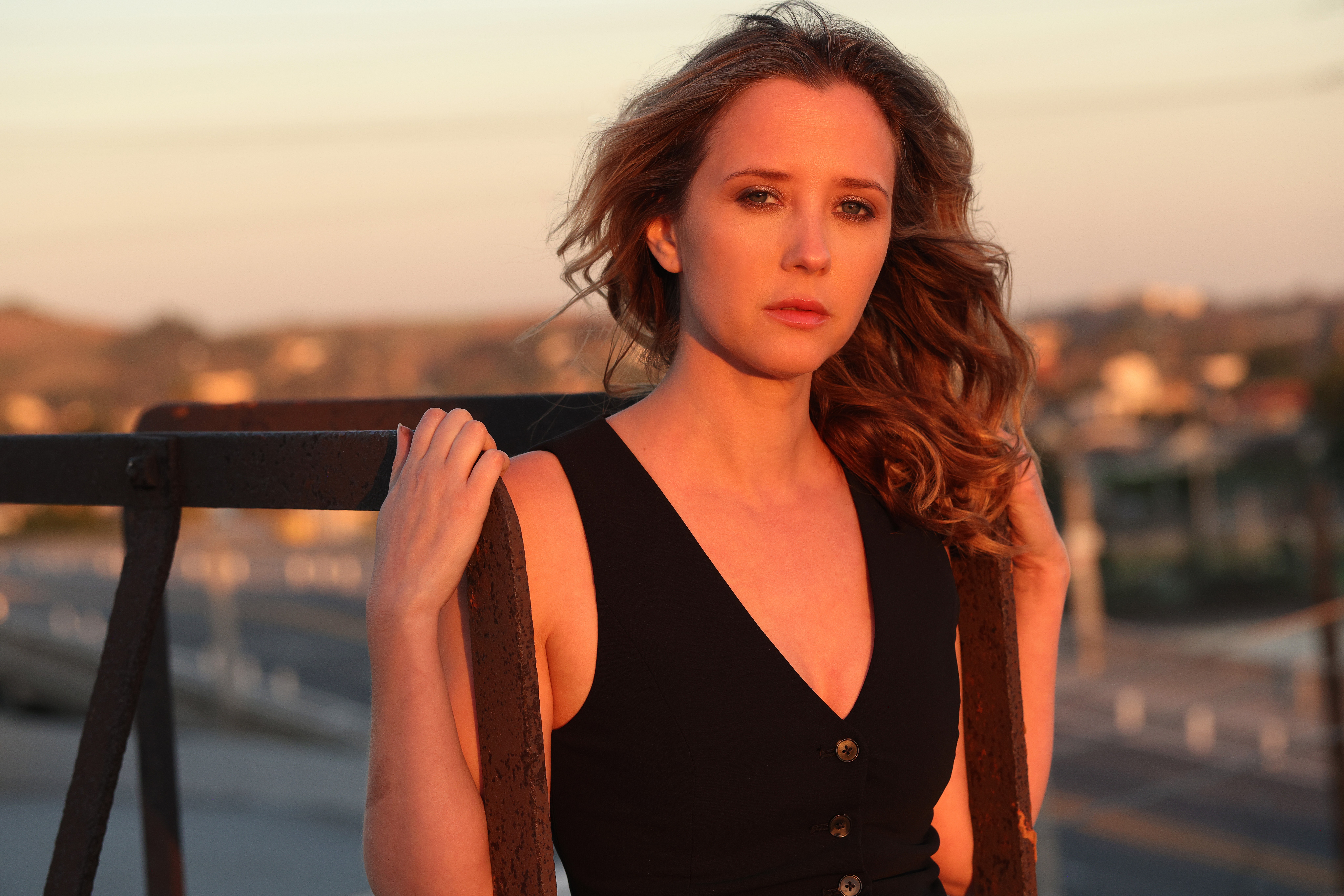 Woman with wavy hair in a black sleeveless top outdoors during sunset, holding a railing, with a cityscape background.