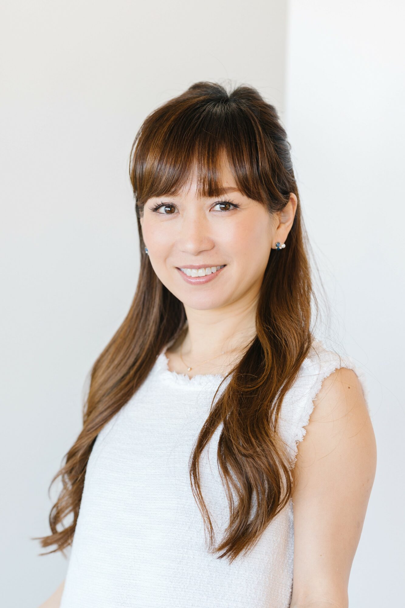 Young woman with long brown hair smiling, wearing a white sleeveless top, standing indoors.