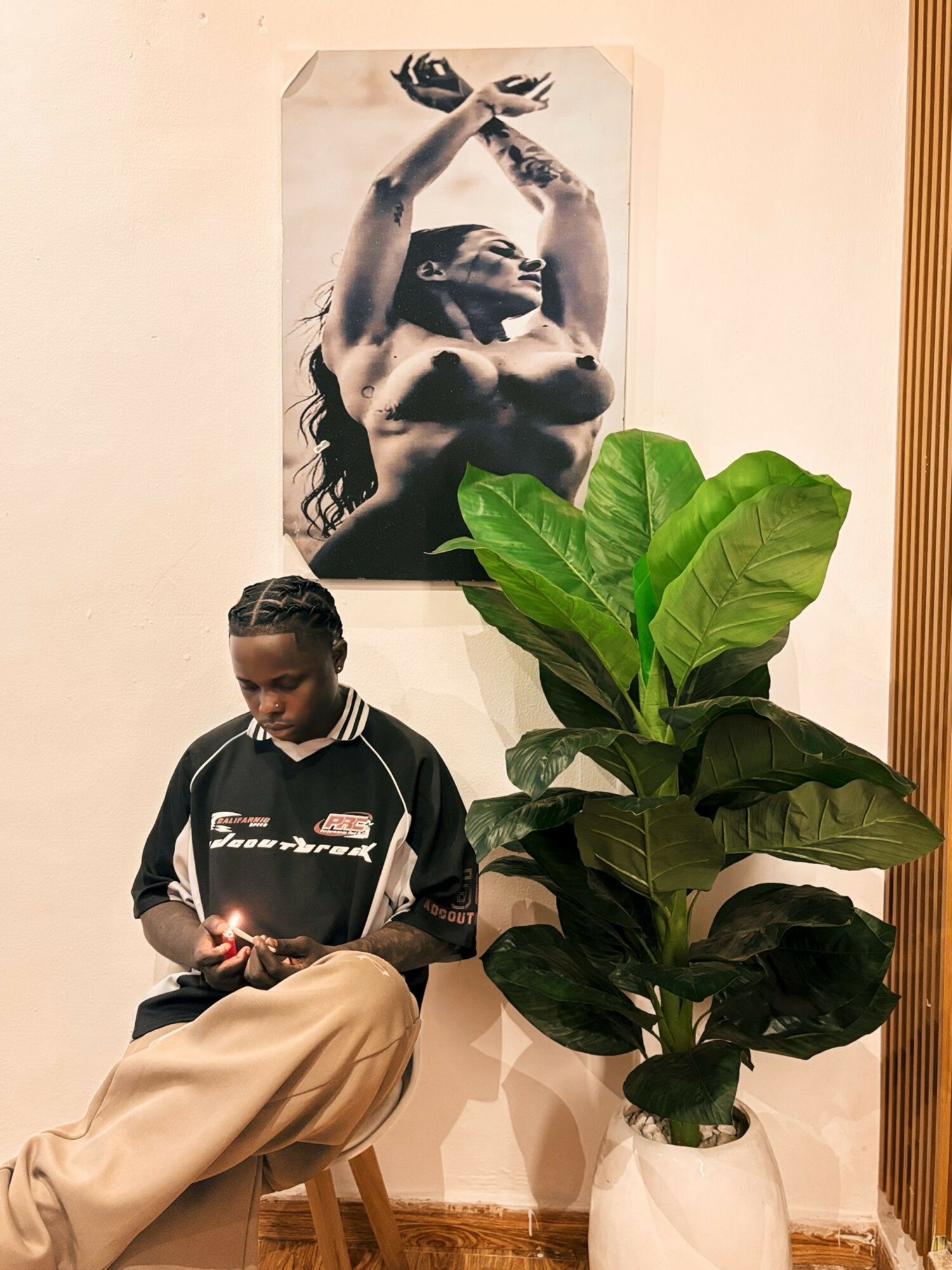 Person sitting on a stool, looking at a phone, with a large green plant nearby and a black-and-white photo of a woman on the wall.