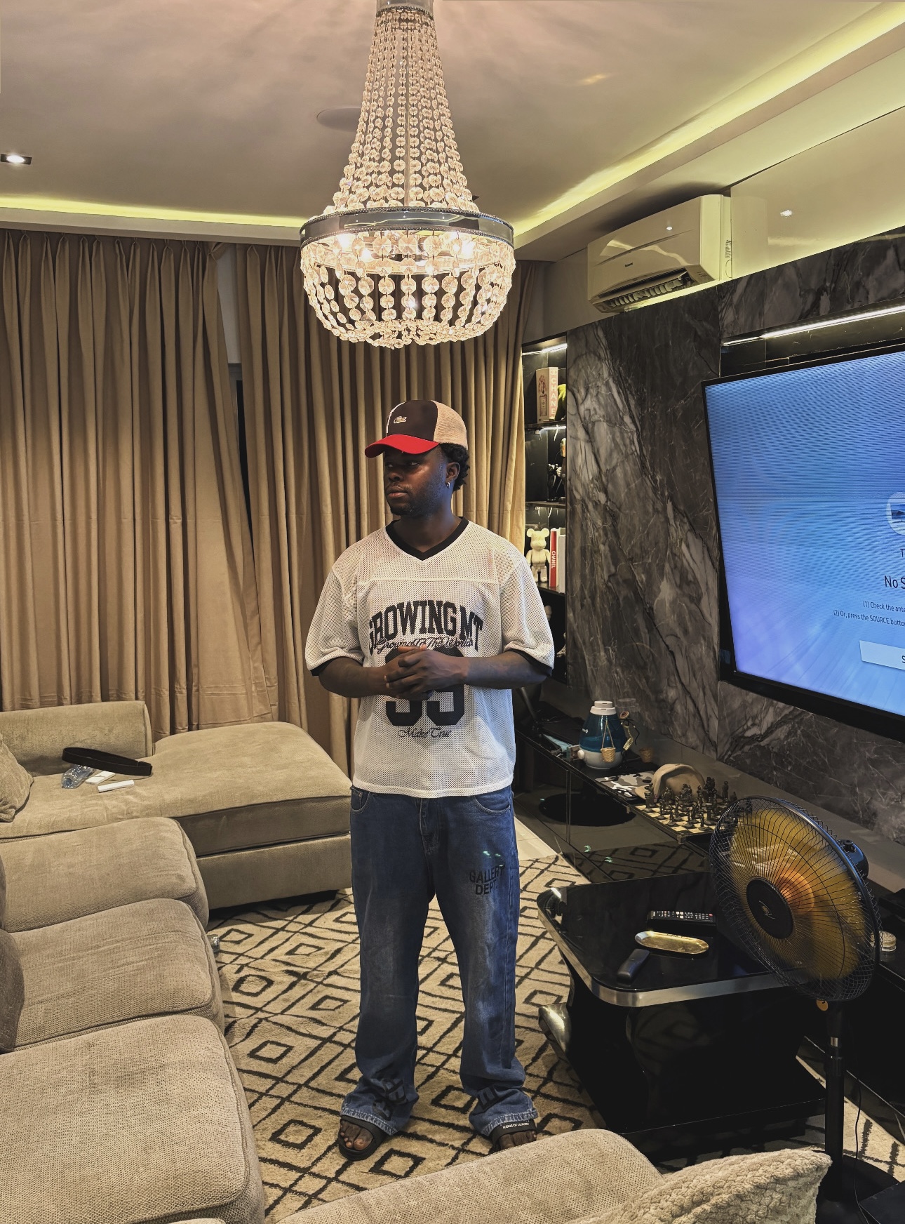 Person wearing a white t-shirt, jeans, and a red cap standing in a living room with a chandelier and a large TV.