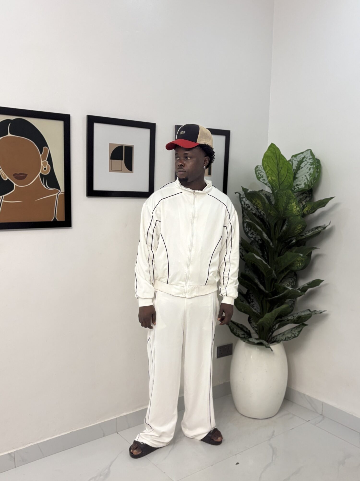 Young man in white tracksuit and cap standing indoors near a potted plant and framed artwork.