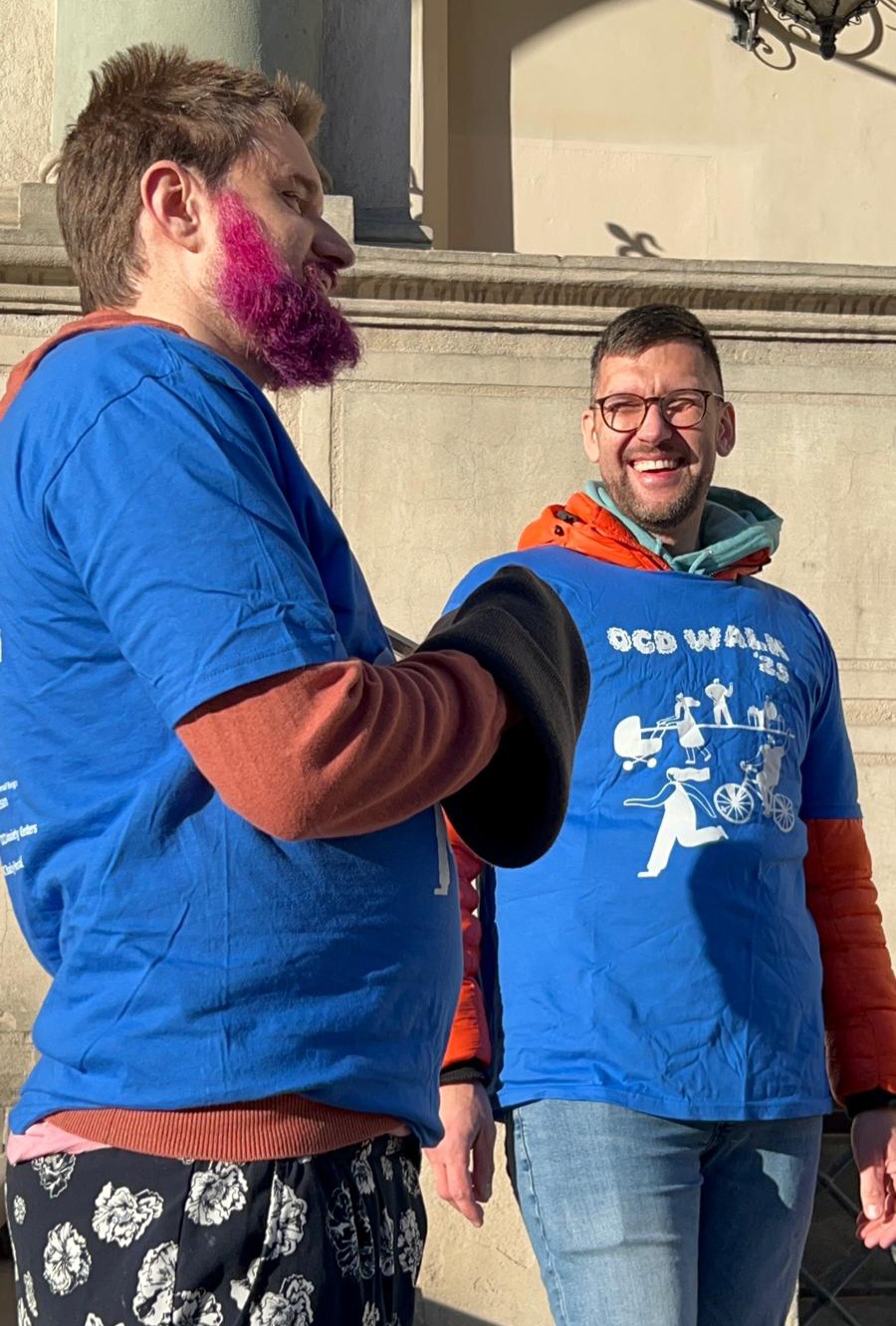 Two men smiling outdoors, one with pink beard and blue shirt, the other with glasses and blue graphic shirt.