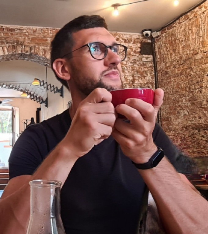 Man with glasses holding a red bowl, sitting indoors with brick walls and a window in the background.