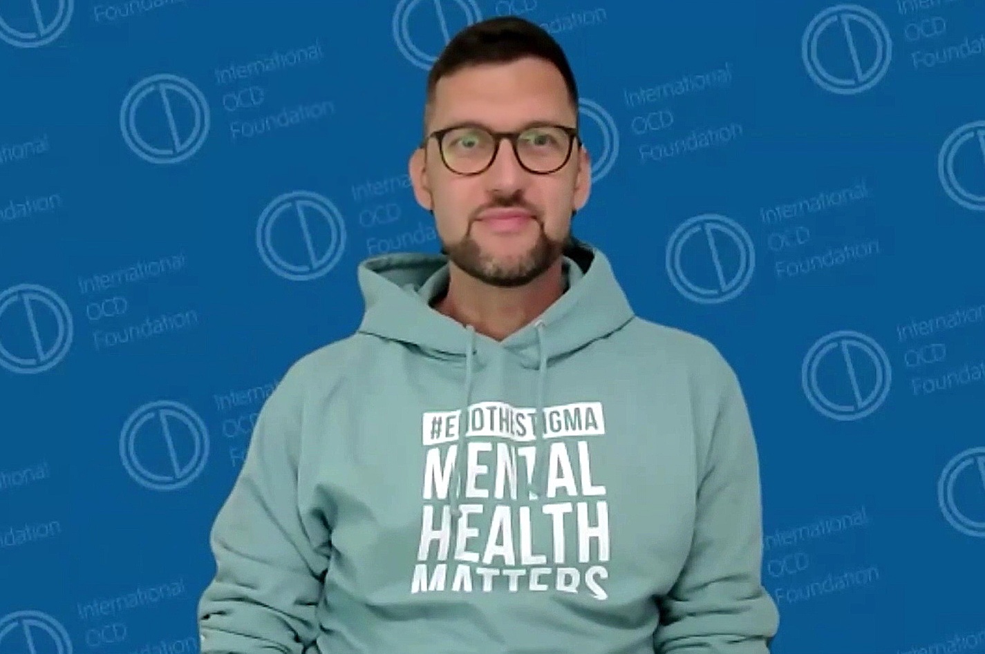 Man with glasses and beard wearing a light green hoodie with text, standing against a blue background.