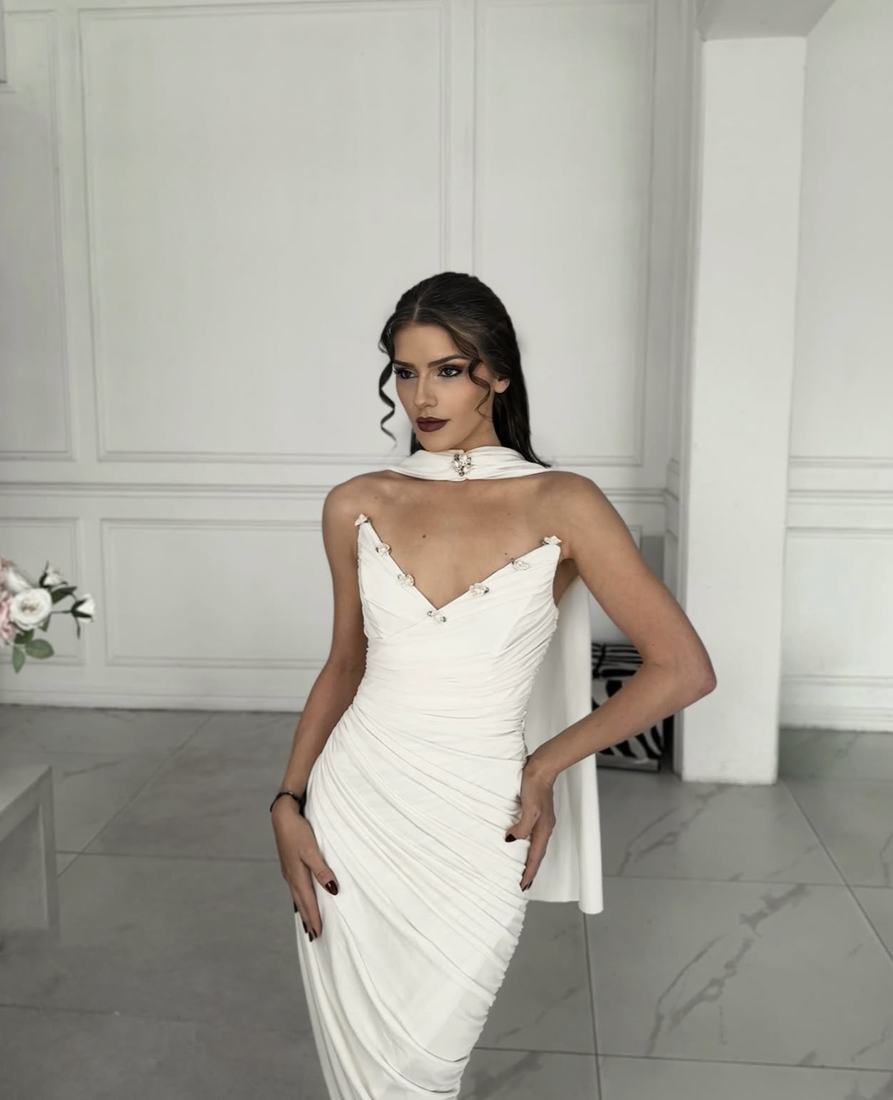 Woman in white dress standing indoors with white paneled walls, posing with hands on hips, looking to the side.