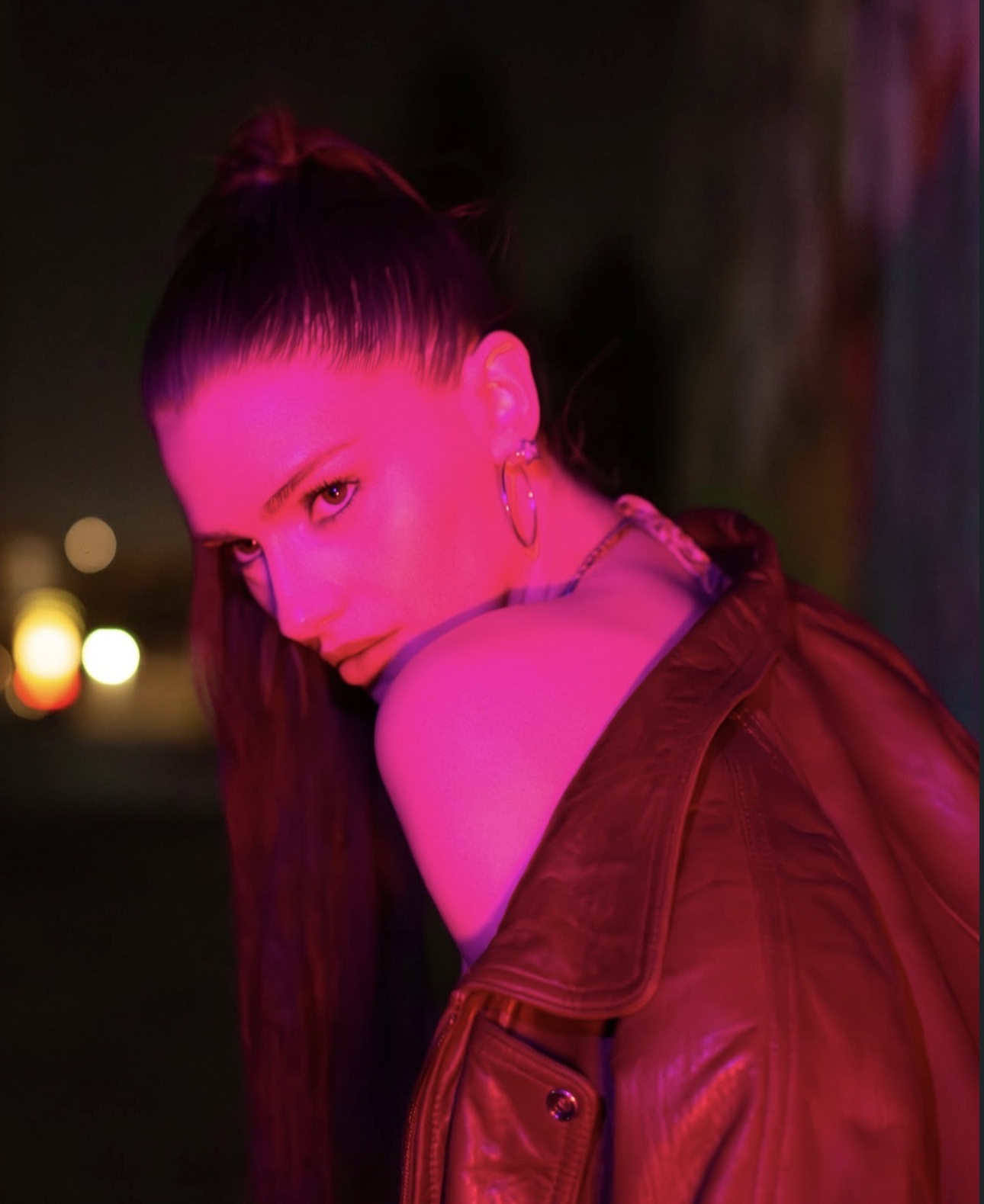 Woman with dark hair in a high ponytail, wearing hoop earrings and a leather jacket, illuminated by pink light.