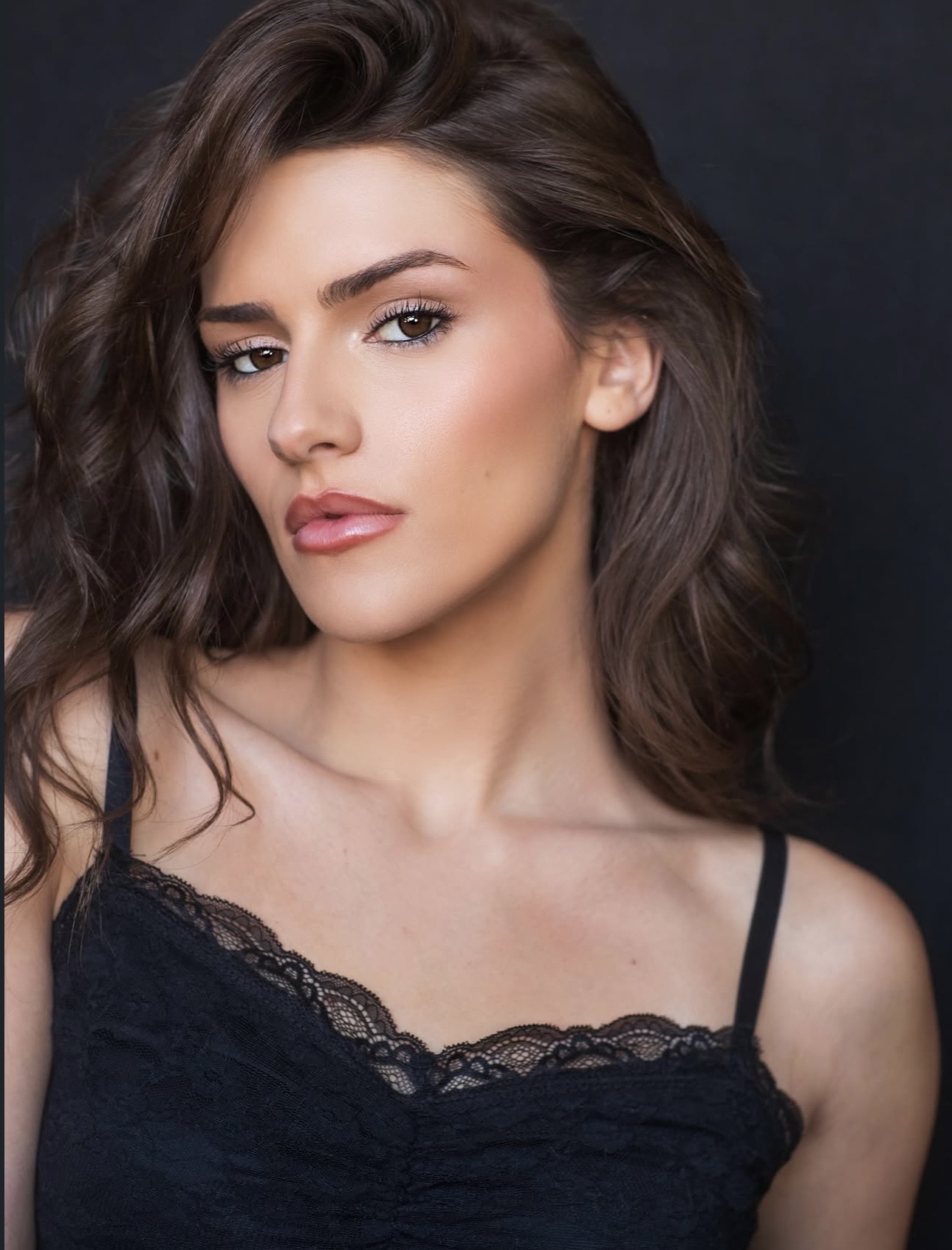 Young woman with wavy brown hair and makeup, wearing a black lace top against a dark background.