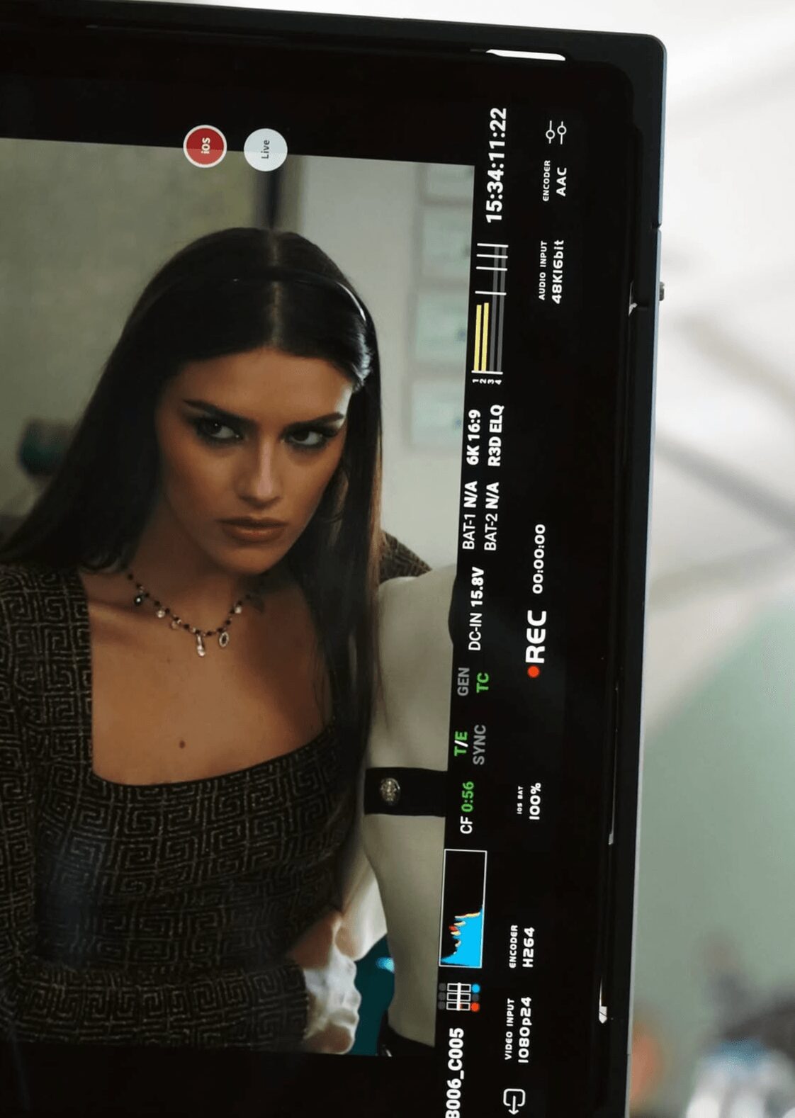 A woman with dark hair and a necklace looks at the camera, with a camera monitor showing her image.