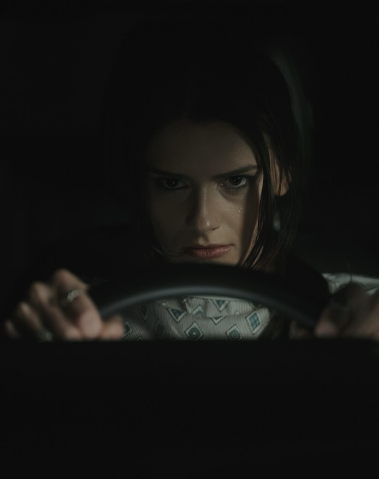 Woman with dark hair looking intensely forward, hands on a steering wheel in a dark setting.