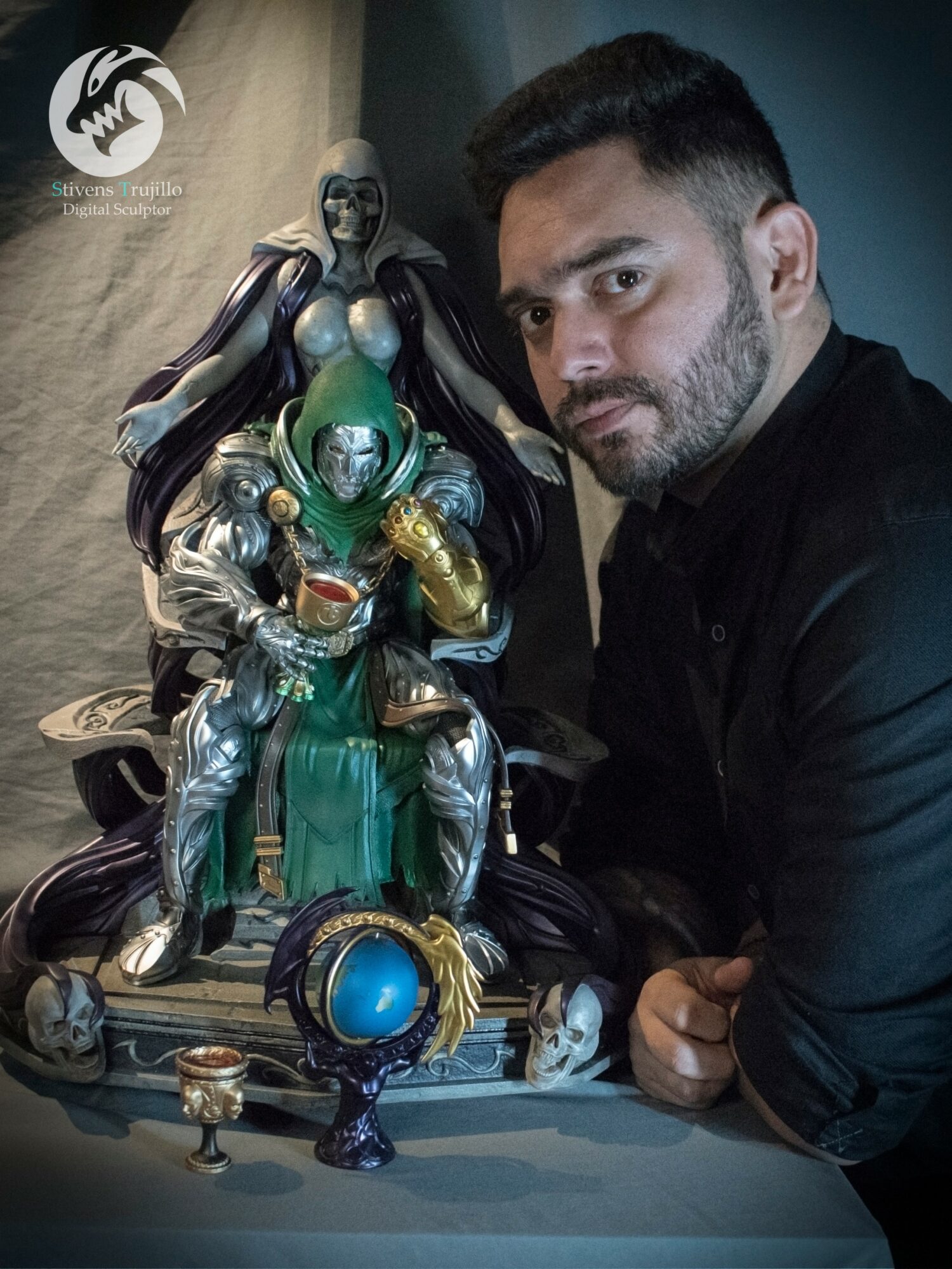 Man with dark hair and beard next to a detailed sculpture of multiple figures, including a green and gold figure, on a table.