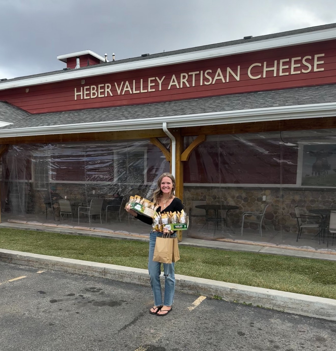 Woman holding cheese and a bag standing outside a cheese shop with a red sign reading 'HEBER VALLEY ARTISAN CHEESE'.