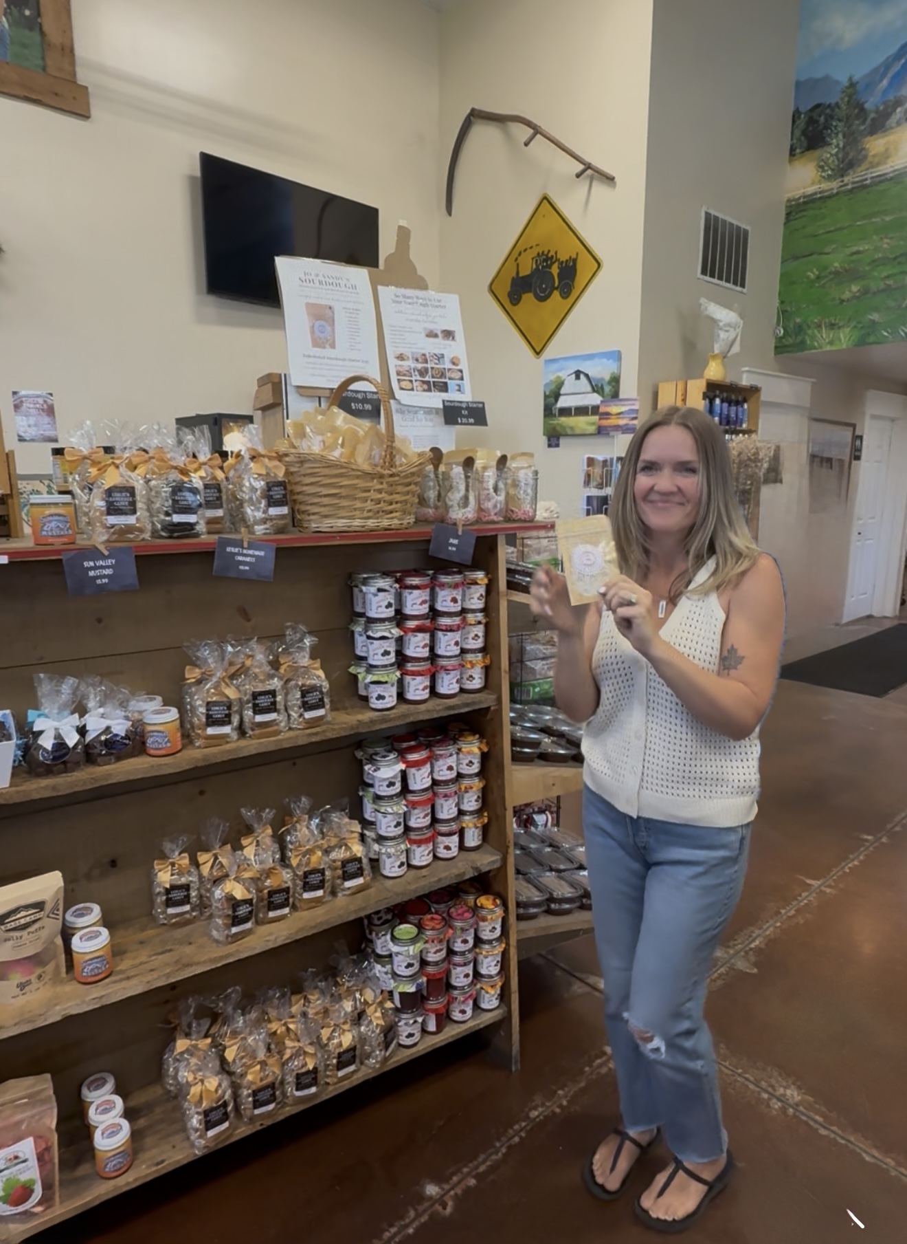 Woman standing next to shelves with jars and baskets, smiling and holding a jar, in a store or market setting.