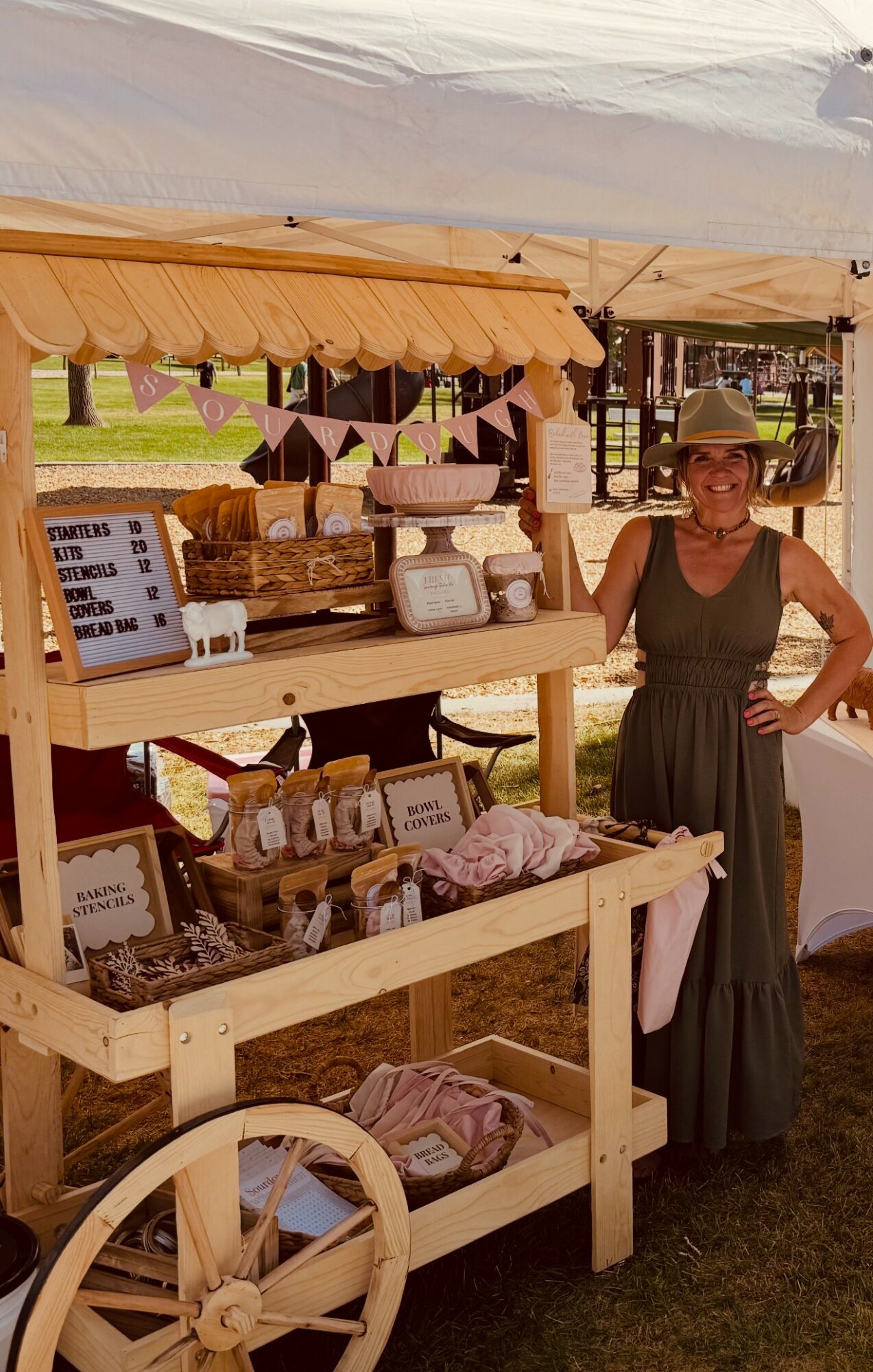 Woman in a hat and dress standing next to a wooden display table with jewelry and accessories at an outdoor market.