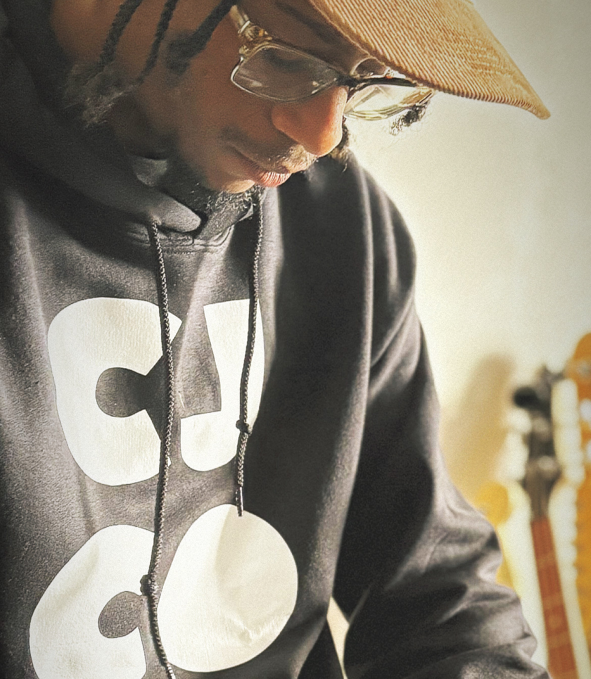 Person wearing a hat, glasses, and a hoodie, looking downward, with a guitar in the background.