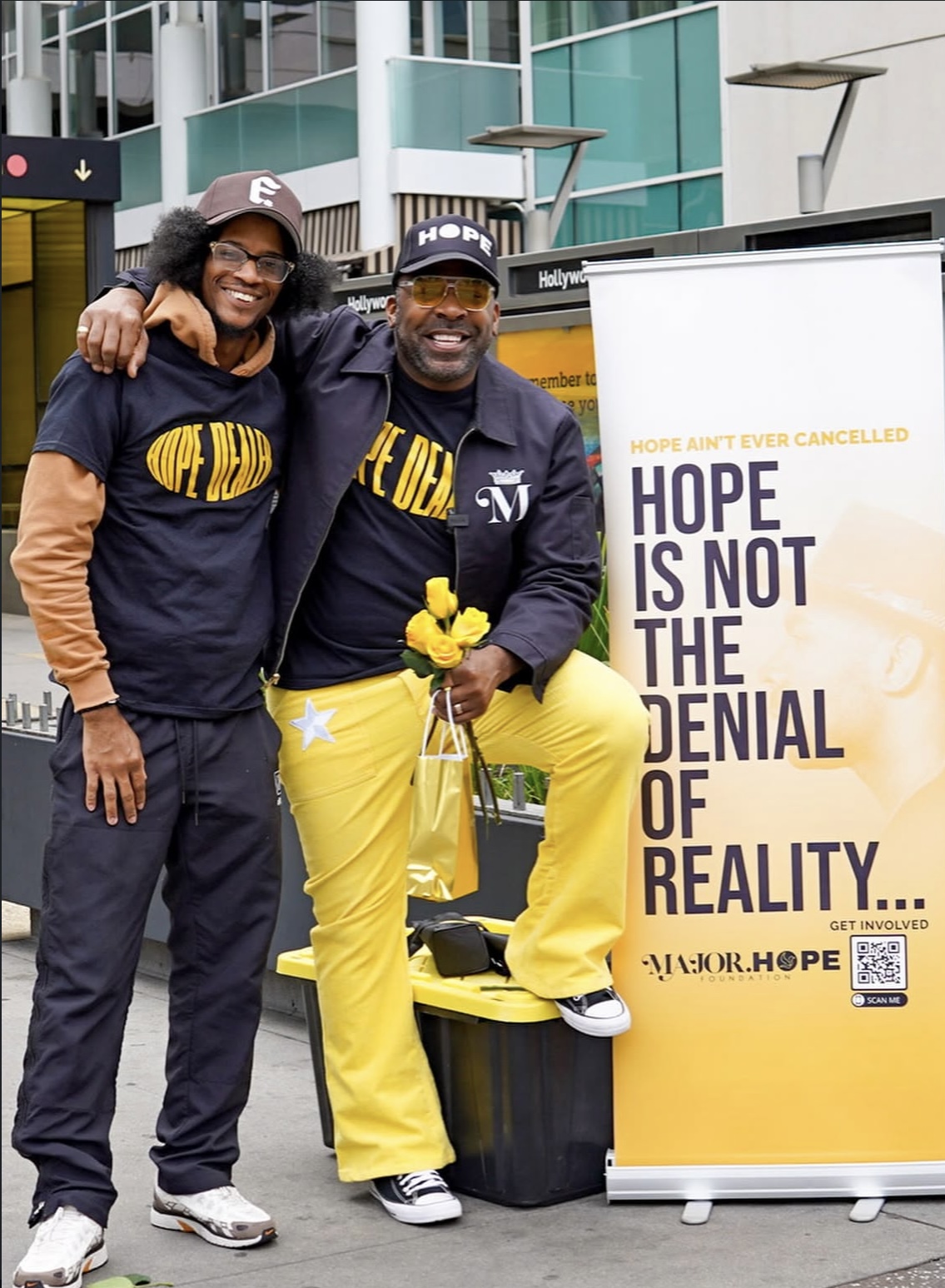 Two people smiling and posing outdoors next to a yellow and white sign about hope and reality.