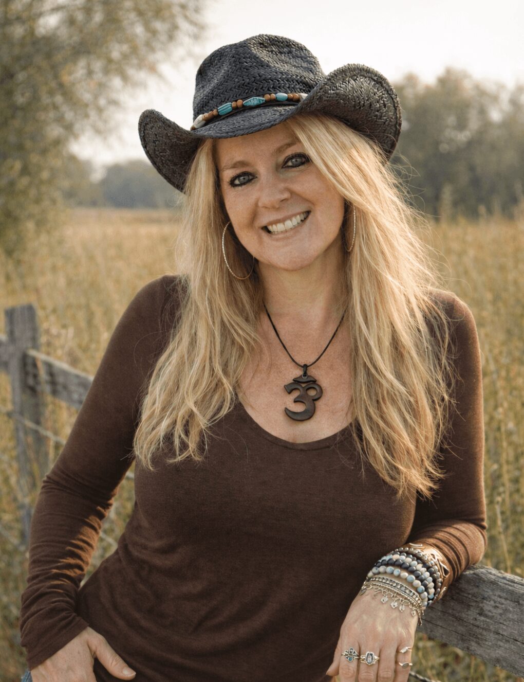 Woman with long blonde hair wearing a black cowboy hat, brown long-sleeve shirt, and jewelry, smiling outdoors.