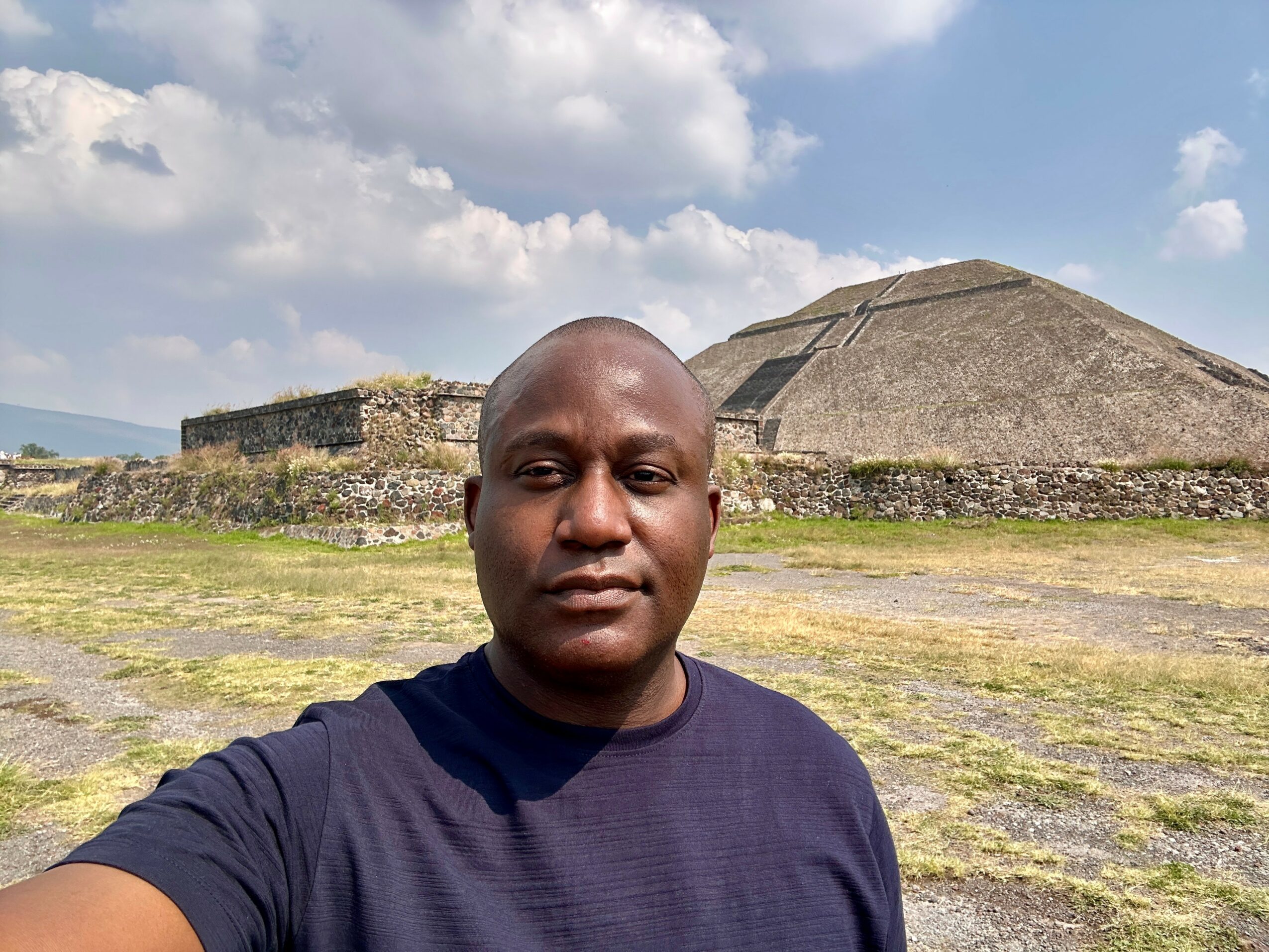 Man standing outdoors with a pyramid-like structure in the background, under a partly cloudy sky.