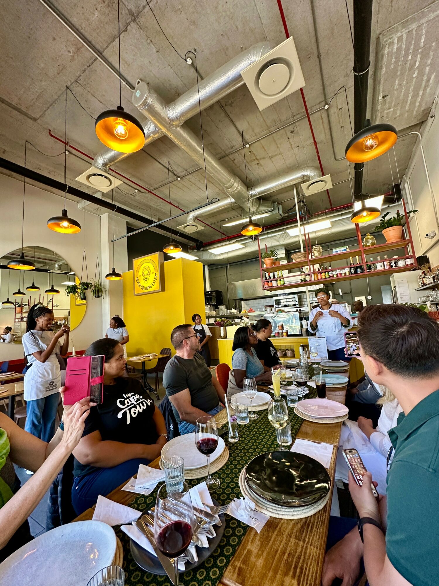 People dining in a modern restaurant with yellow and gray decor, hanging lights, and shelves with bottles, some using phones.