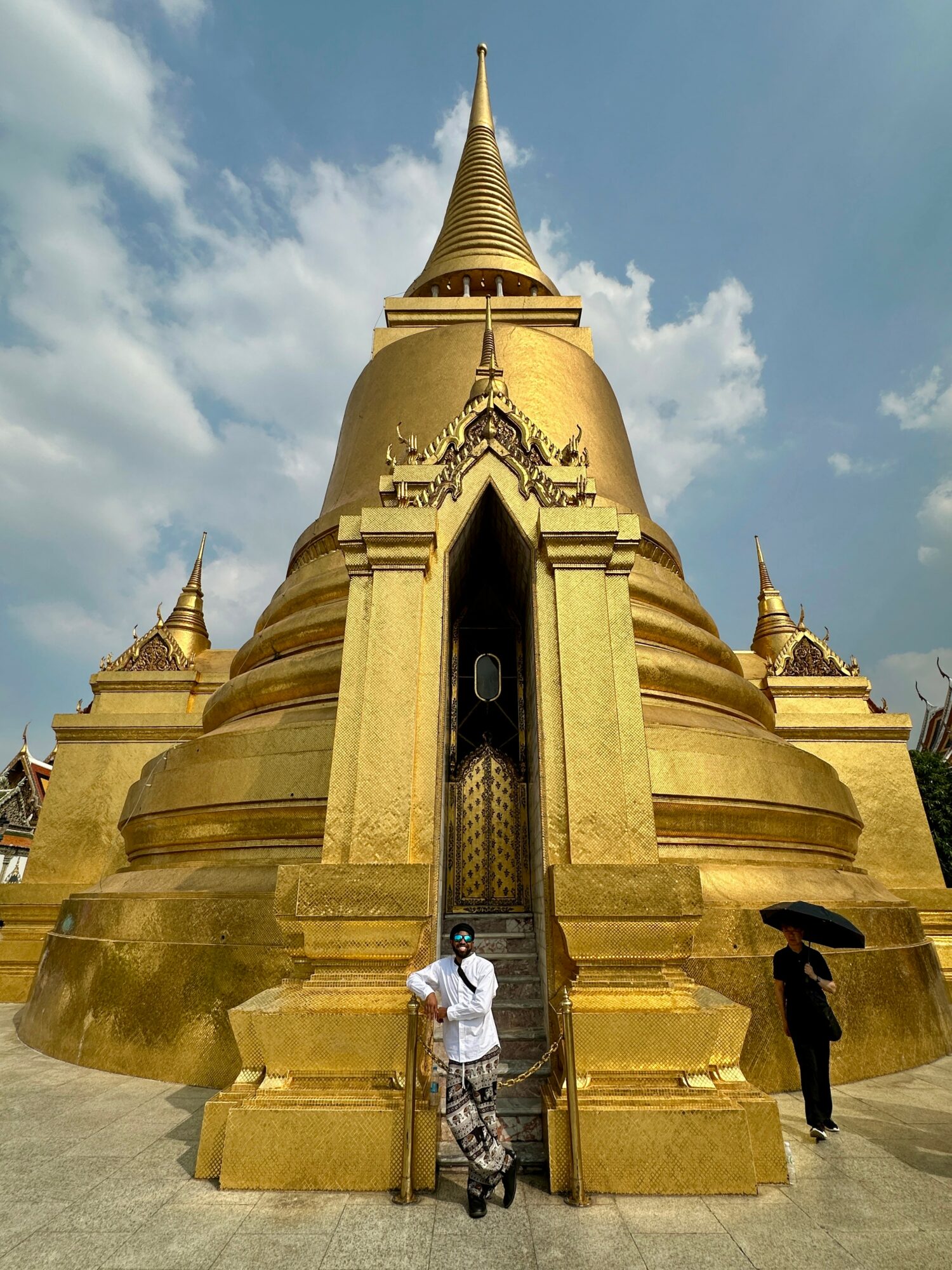 Golden temple with a tall spire, two people standing at the entrance, one holding an umbrella, blue sky with clouds