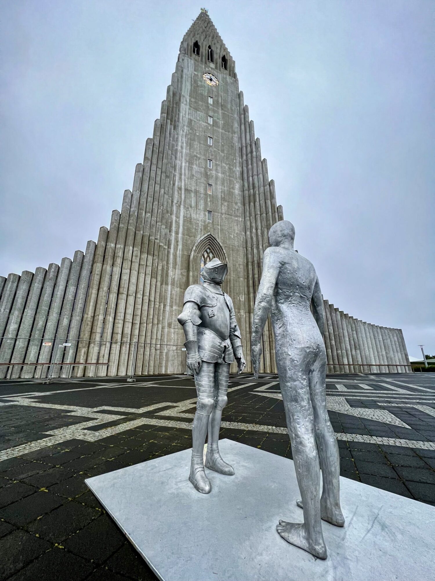 Statues of a child and an adult holding hands in front of a tall, pointed building with vertical ridges.