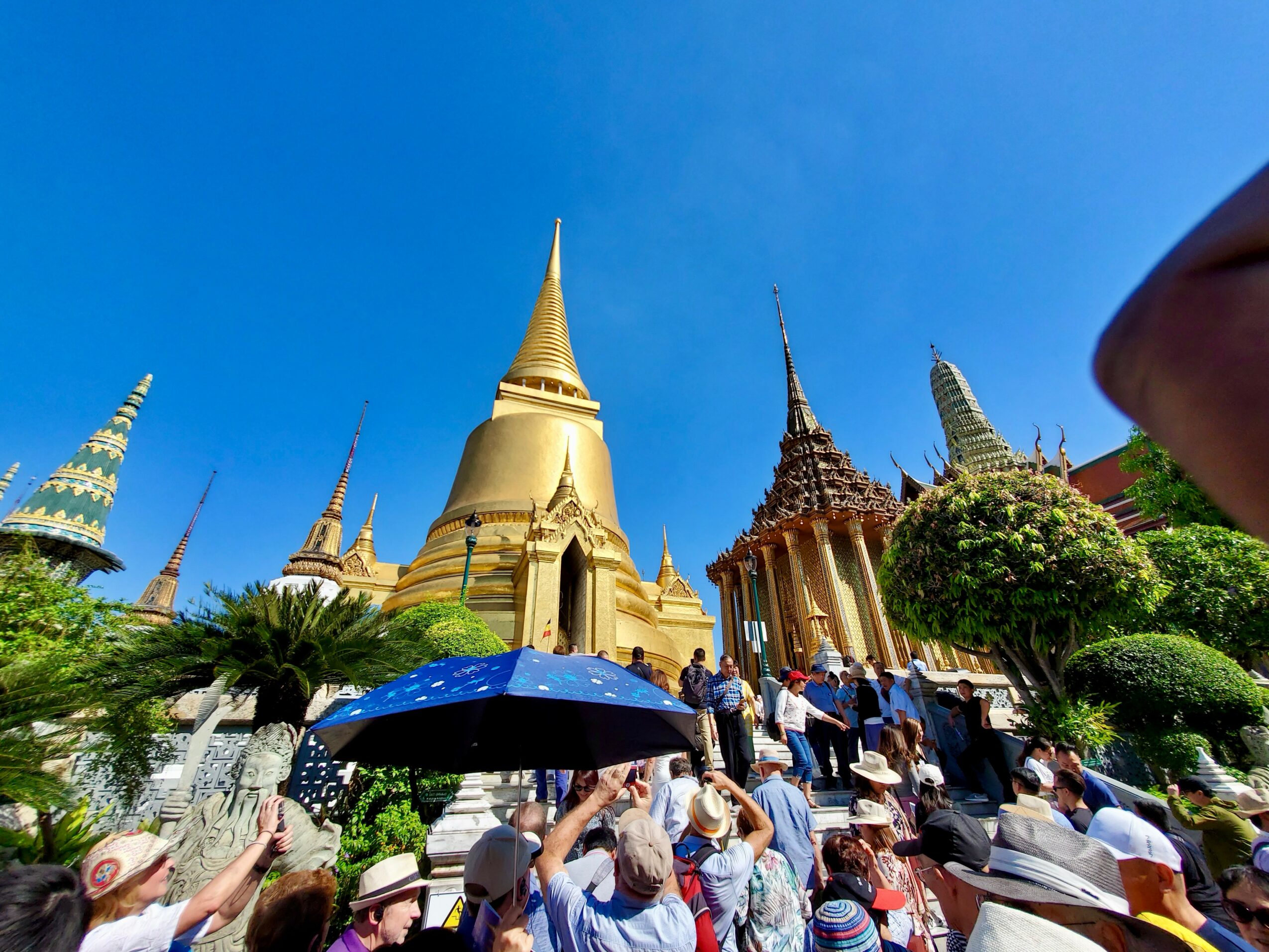 People gather at a temple complex with golden spires and ornate structures, some holding umbrellas, under a clear blue sky.
