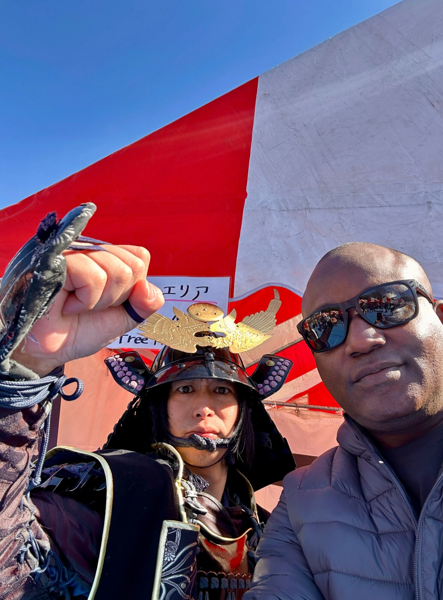 Two people taking a selfie outdoors with a large red and white structure in the background, one wearing sunglasses and the other holding a small crocodile.