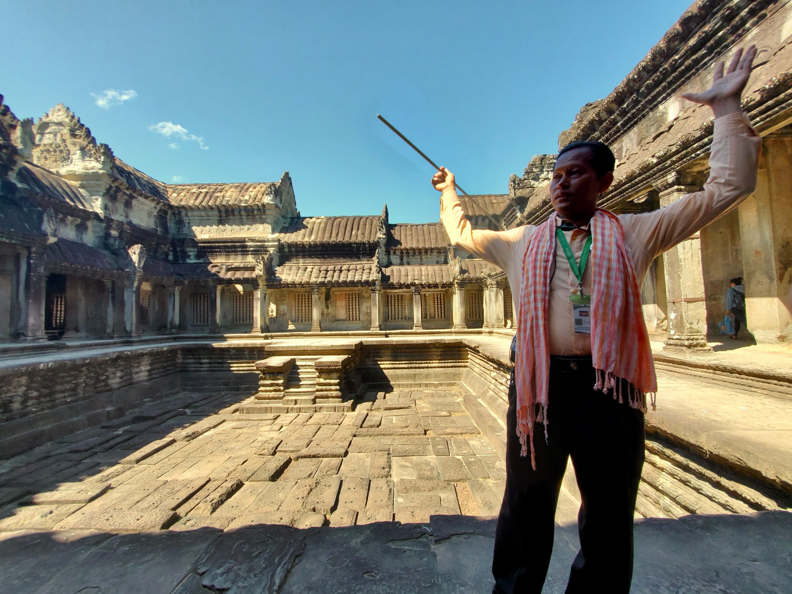 Man standing with arms raised and holding a stick in an ancient stone courtyard with buildings and a clear blue sky.
