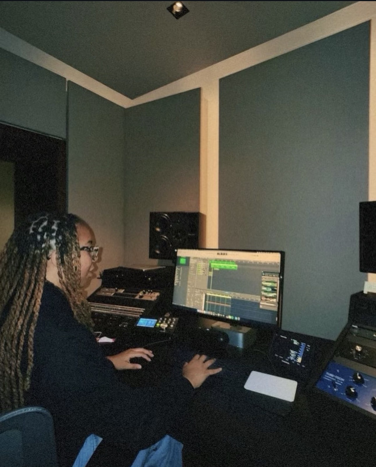 Person with braided hair working at a music production computer setup in a room with blue walls.