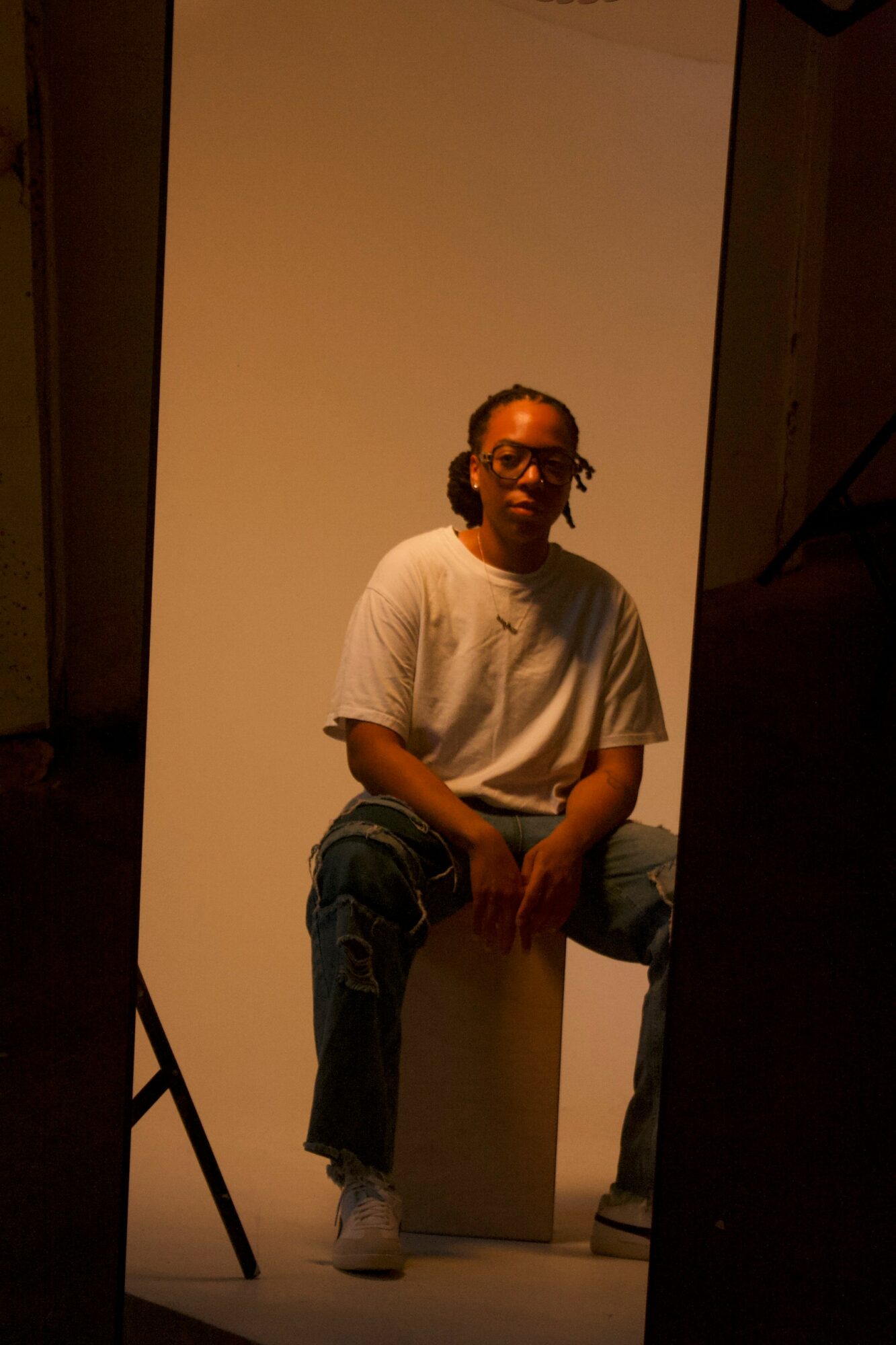 Person sitting on a box in front of a plain background, wearing glasses, a white t-shirt, and jeans.