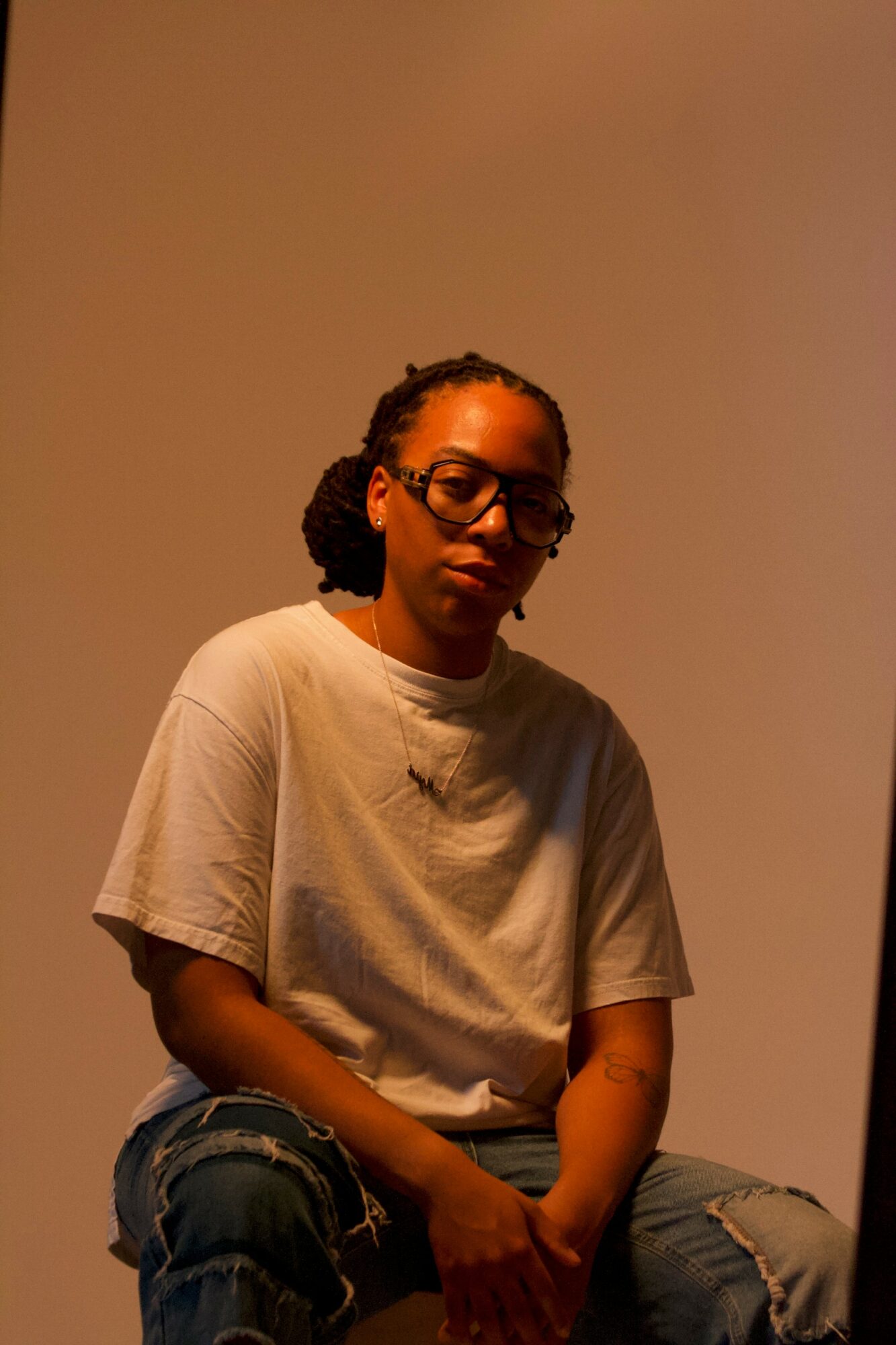 Person with glasses and curly hair sitting against a plain background, wearing a white t-shirt and ripped jeans.