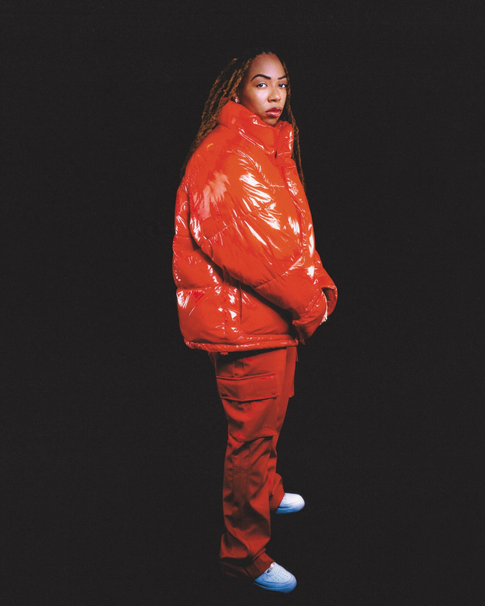 Person with long hair wearing a shiny red puffer jacket and red pants, standing against a black background.