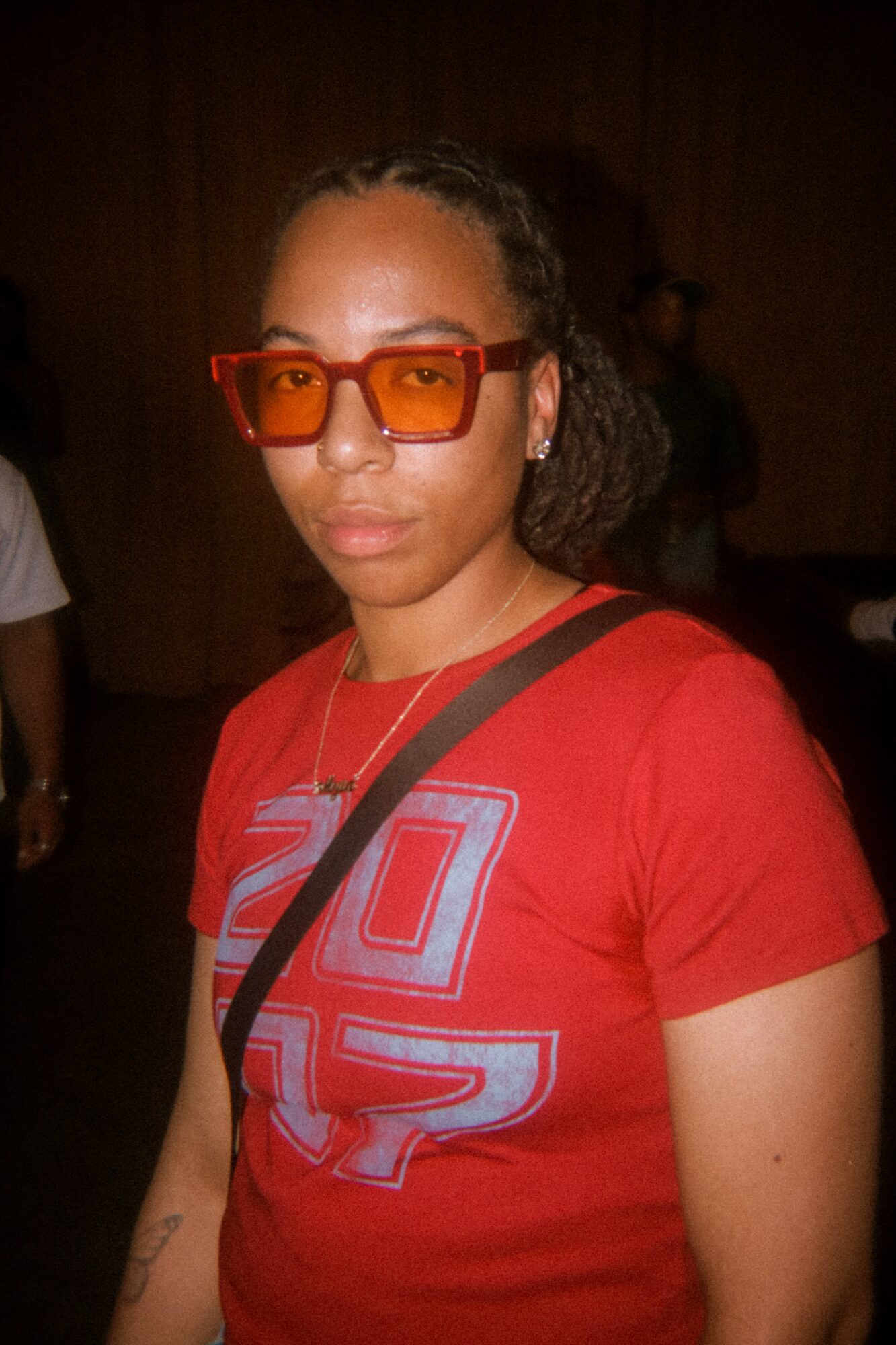 Woman wearing orange sunglasses and a red T-shirt with a graphic design, carrying a black strap across her chest.
