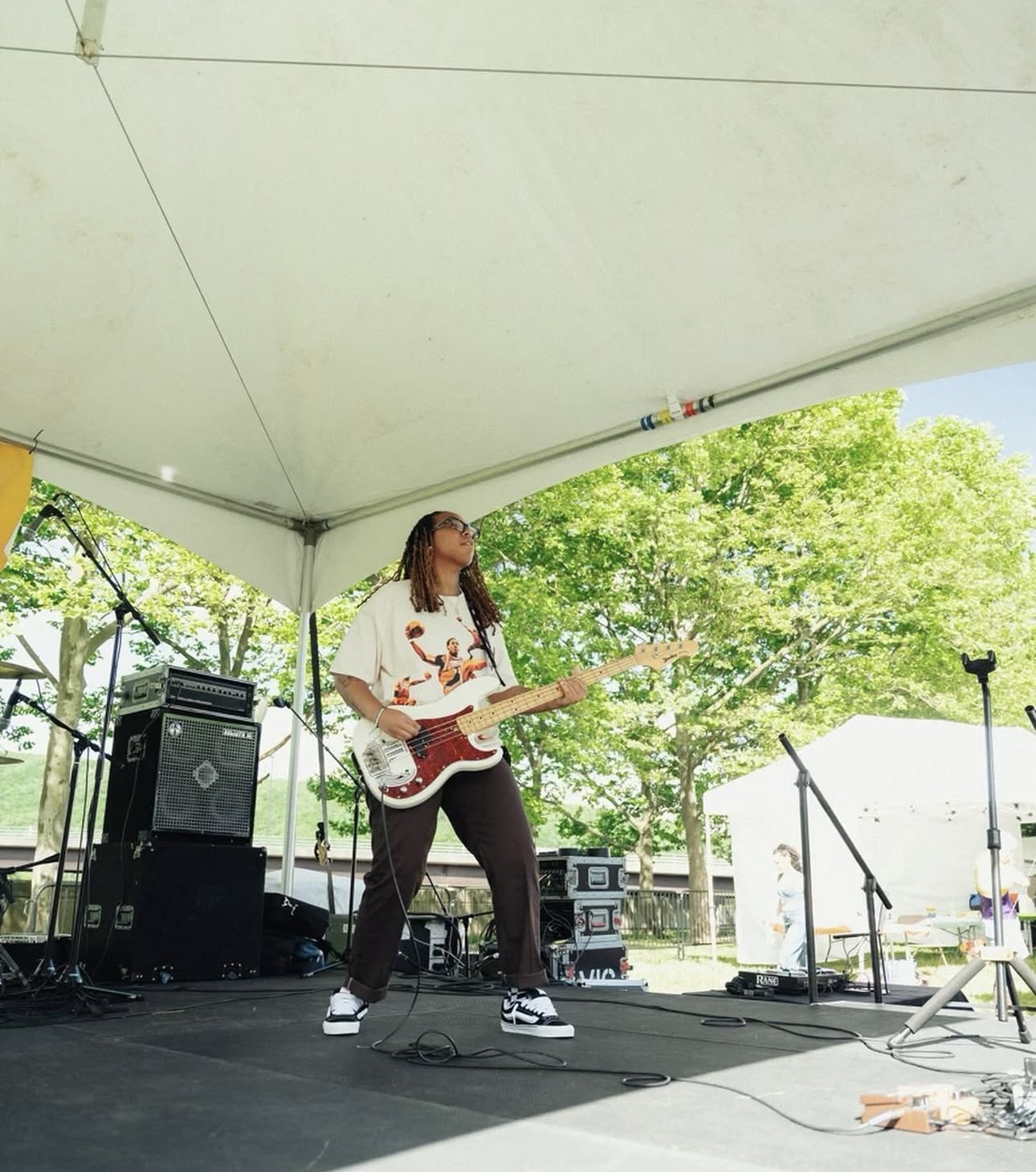 A woman plays an electric guitar on an outdoor stage under a tent, with trees and a building in the background.
